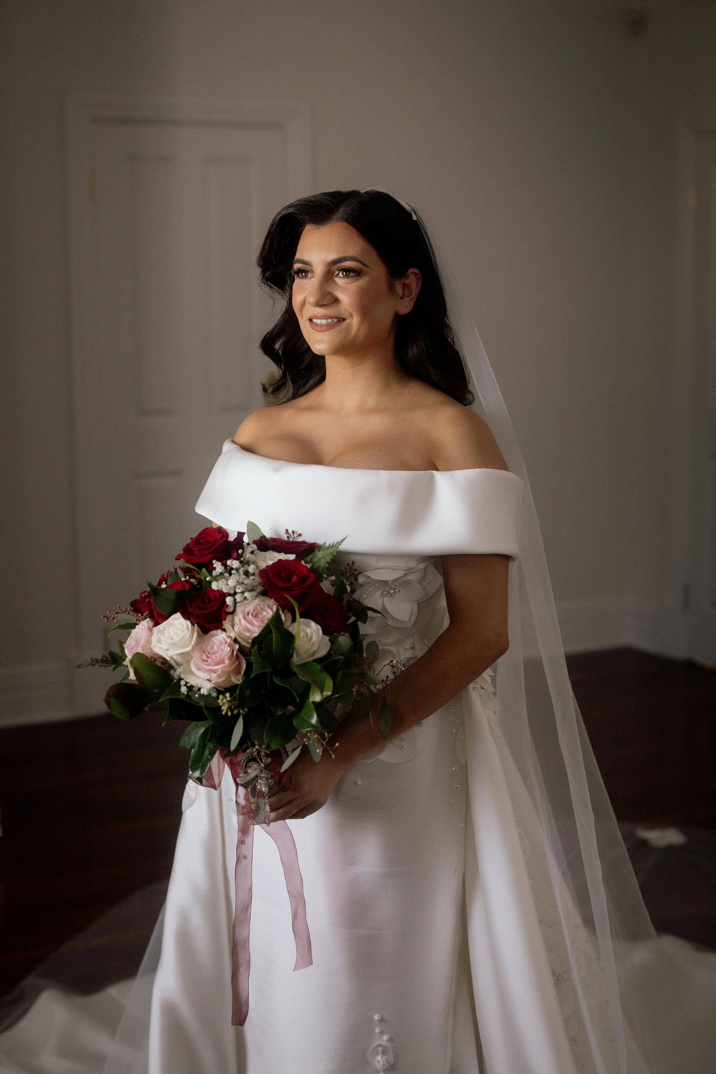 A bride in an off-shoulder satin gown holds a lush bouquet of red and pink roses, captured in a cinematic bridal portrait.