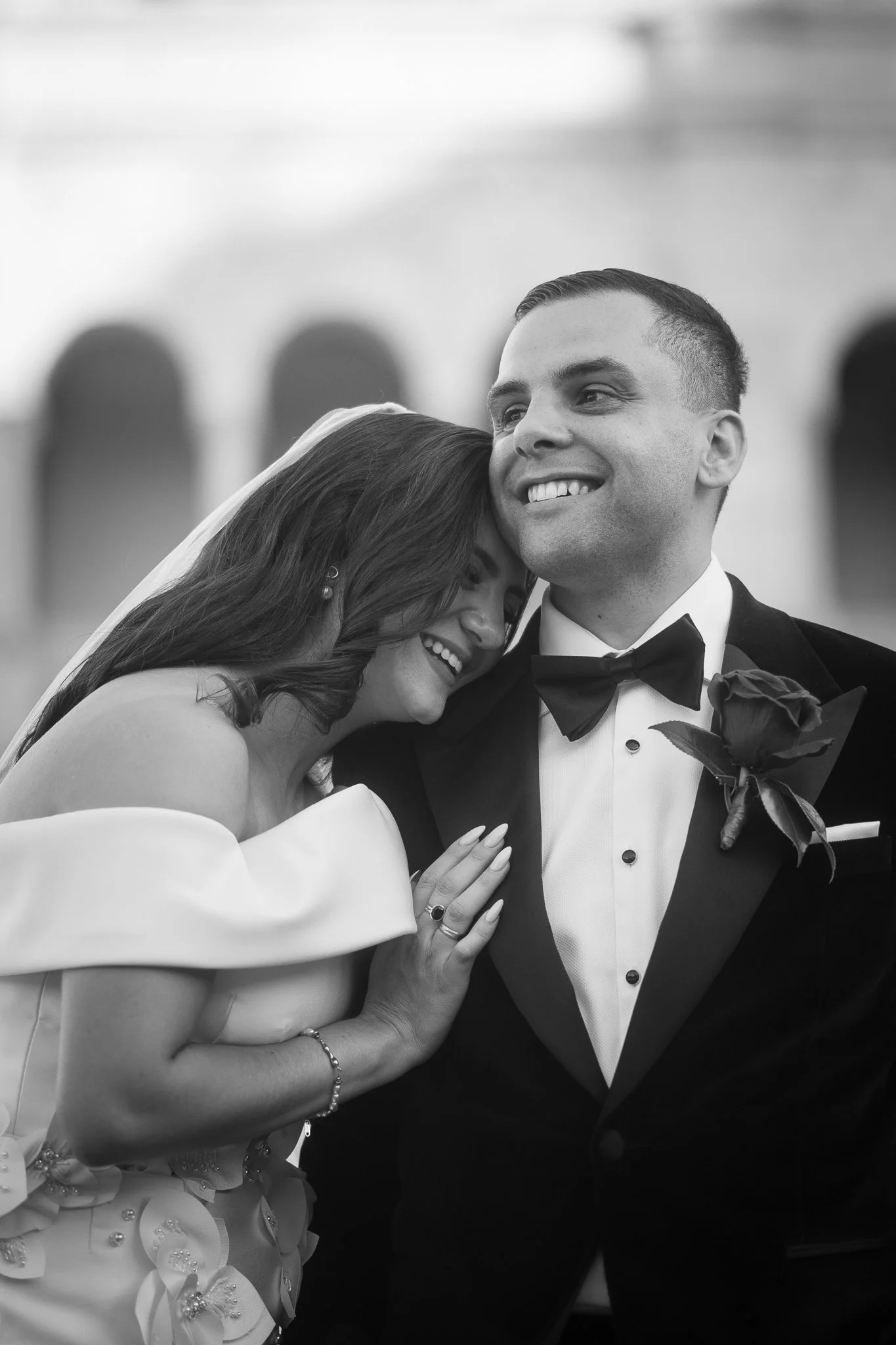 Laughing bride in off-shoulder gown rests her head on a smiling groom's shoulder in a candid wedding portrait.