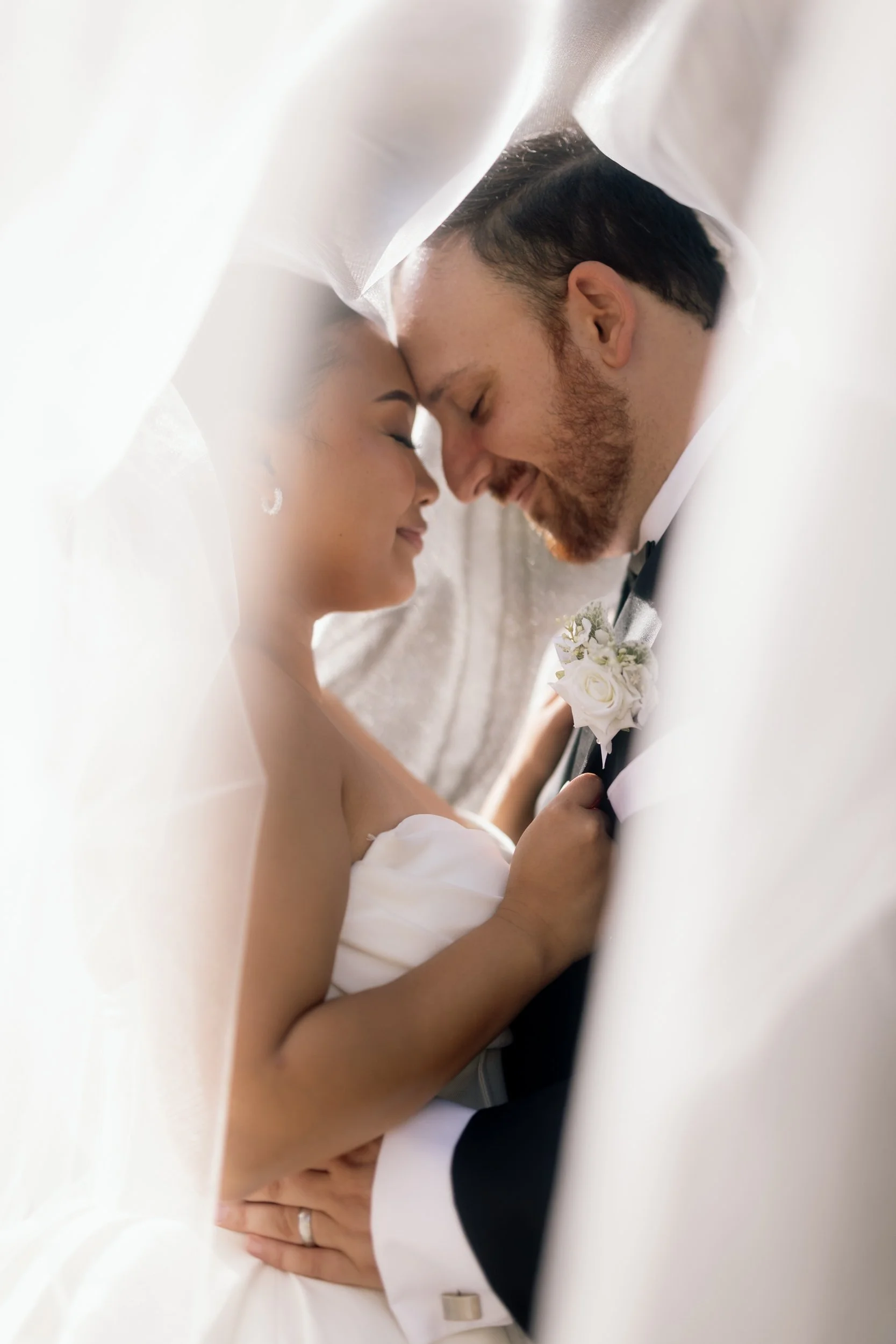 Bride and groom share a tender forehead touch framed by her flowing veil, his white rose boutonniere caught in warm light.