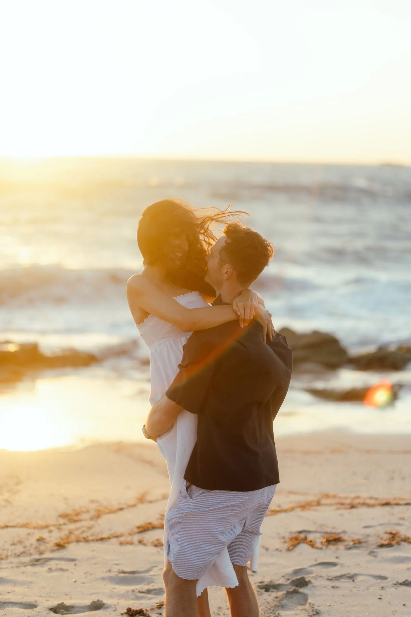 A groom lifts his bride on a sun-drenched beach at golden hour, waves softly breaking behind them during their pre-wedding shoot