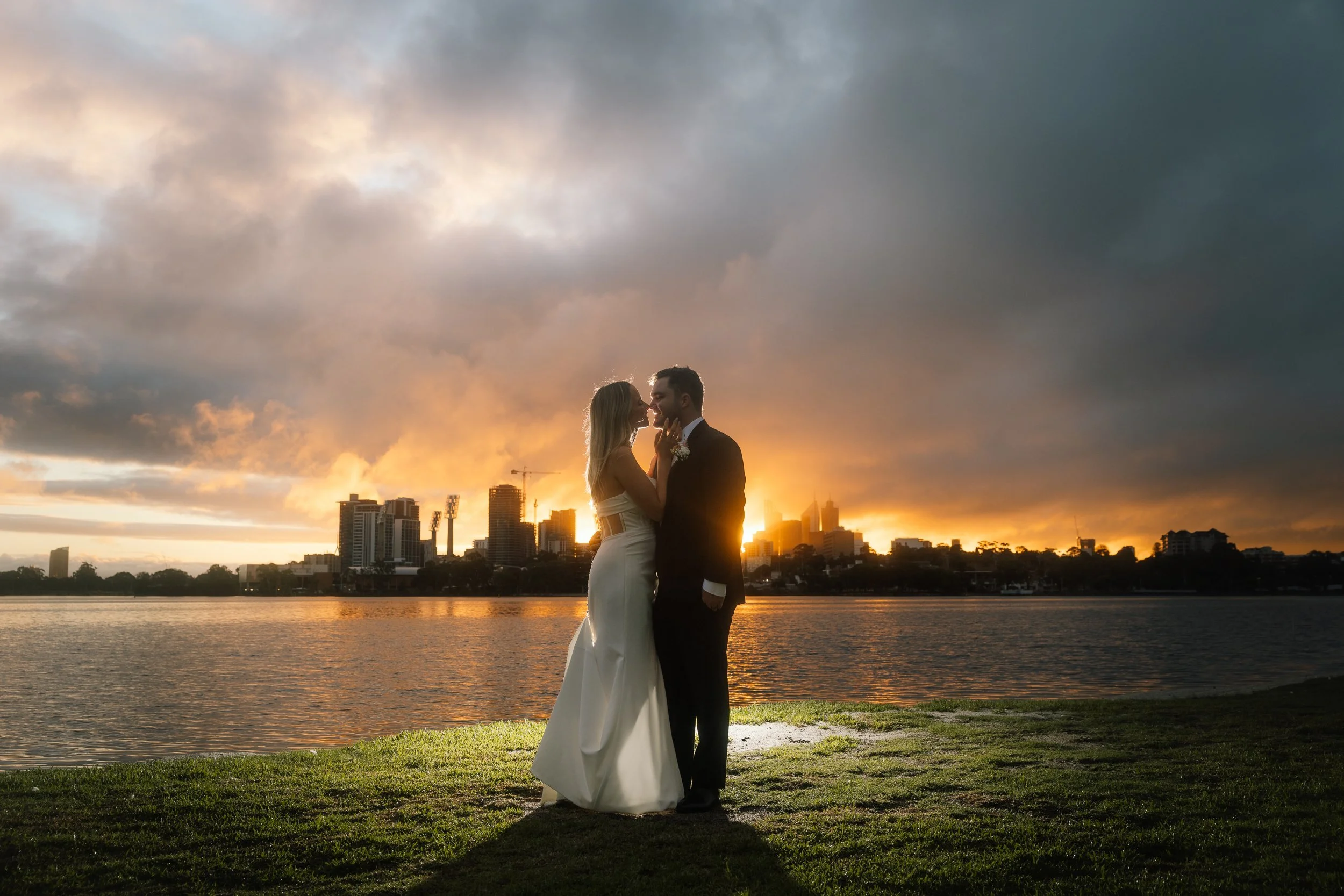 Bride in sleek white gown and groom in black suit share a tender moment by the Swan River as a fiery sunset silhouettes