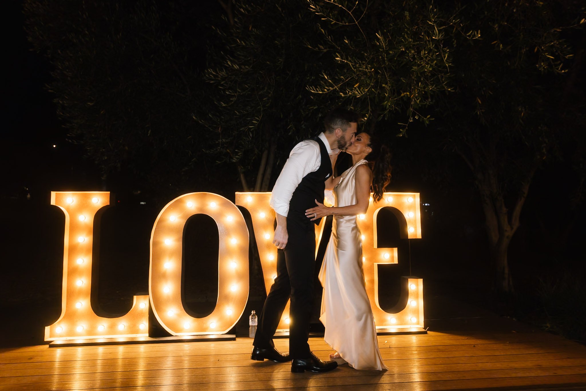 Bride in a champagne satin gown and groom kissing in front of large illuminated LOVE marquee letters at night at The Mallard Duck Swan Valley Perth, photographed by James Di Stefano