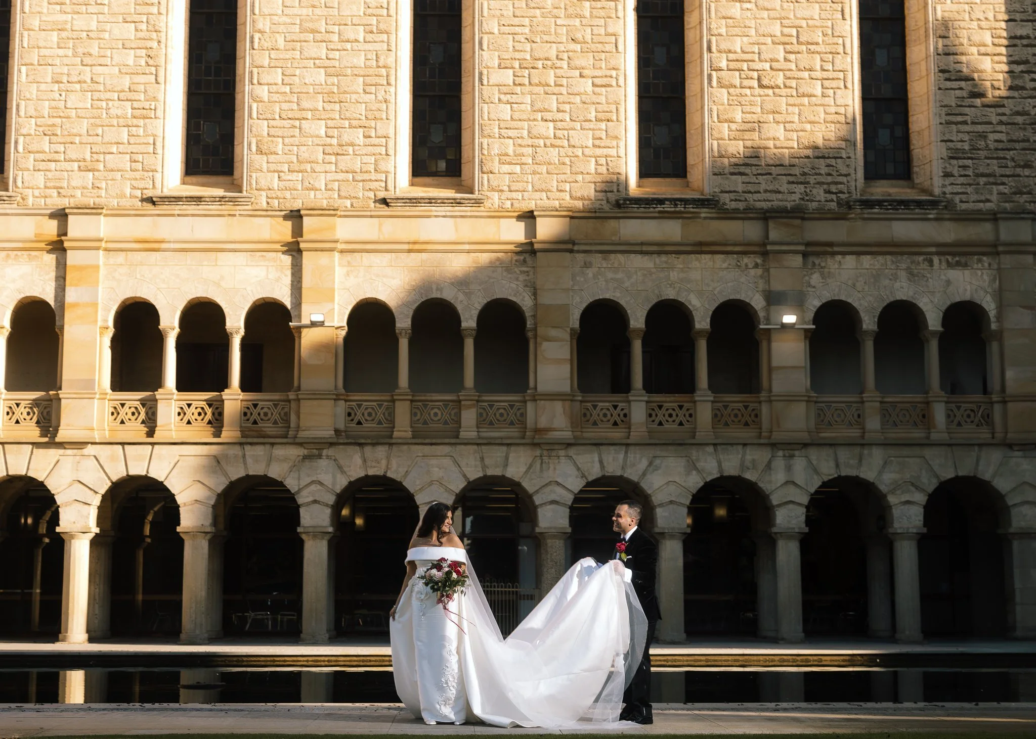 Bride in an off-shoulder gown holds a red bouquet as her groom lifts her train before UWA's arched limestone cloisters.