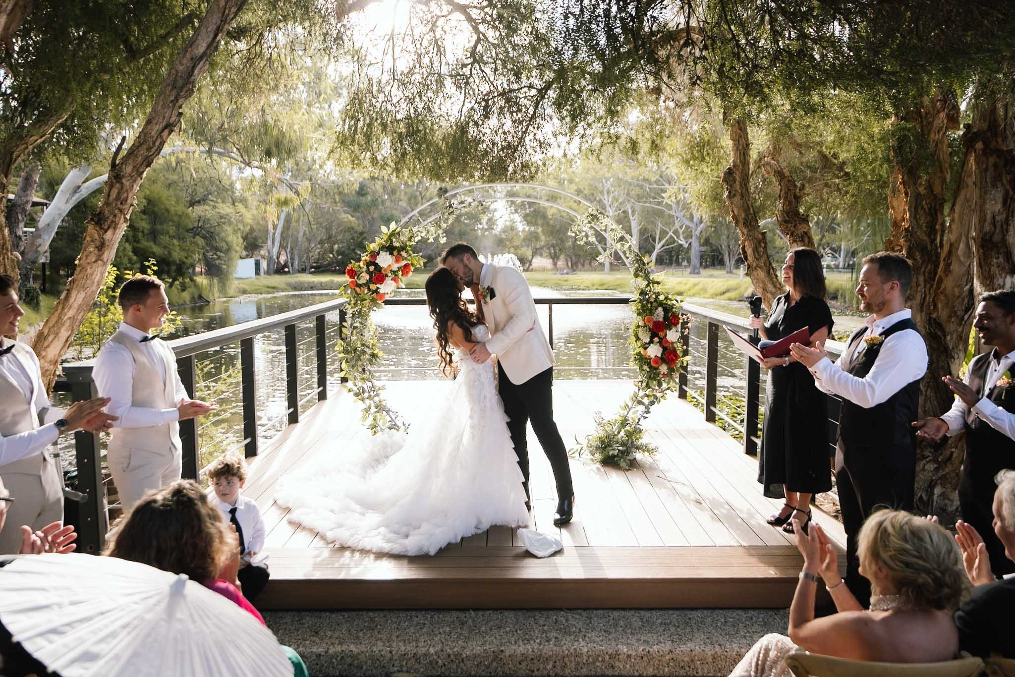 Bride and groom sharing their first kiss on the lakeside timber deck at The Mallard Duck Swan Valley Perth, framed by a colourful floral arch and surrounding native trees, photographed by James Di Stefano