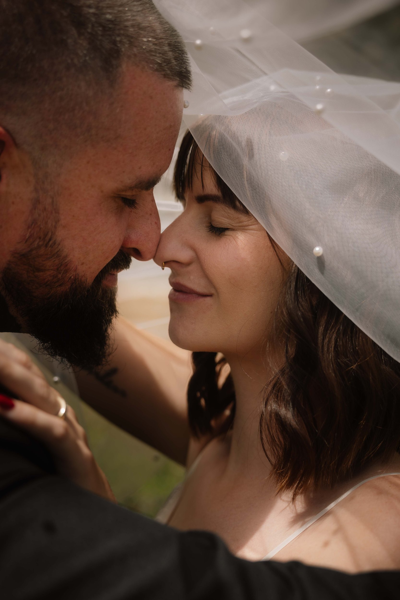 Bride and groom share a tender nose-to-nose moment beneath a pearl-trimmed wedding veil, eyes softly closed.