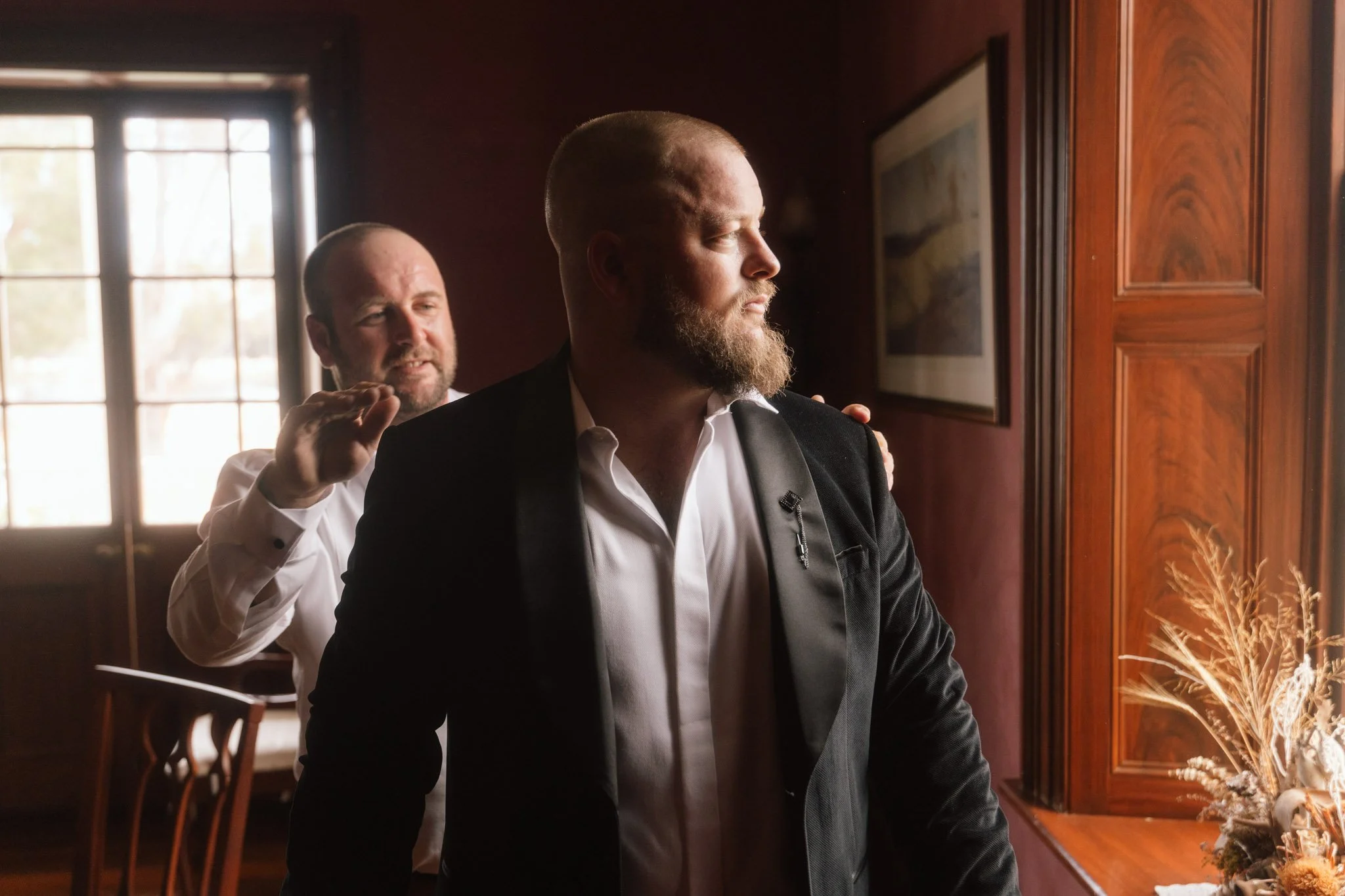 A groom in a black velvet wedding suit looking out a historic wooden window while his groomsman assists him, captured in moody directional side light inside the heritage manor house at Buckland Estate, Western Australia.