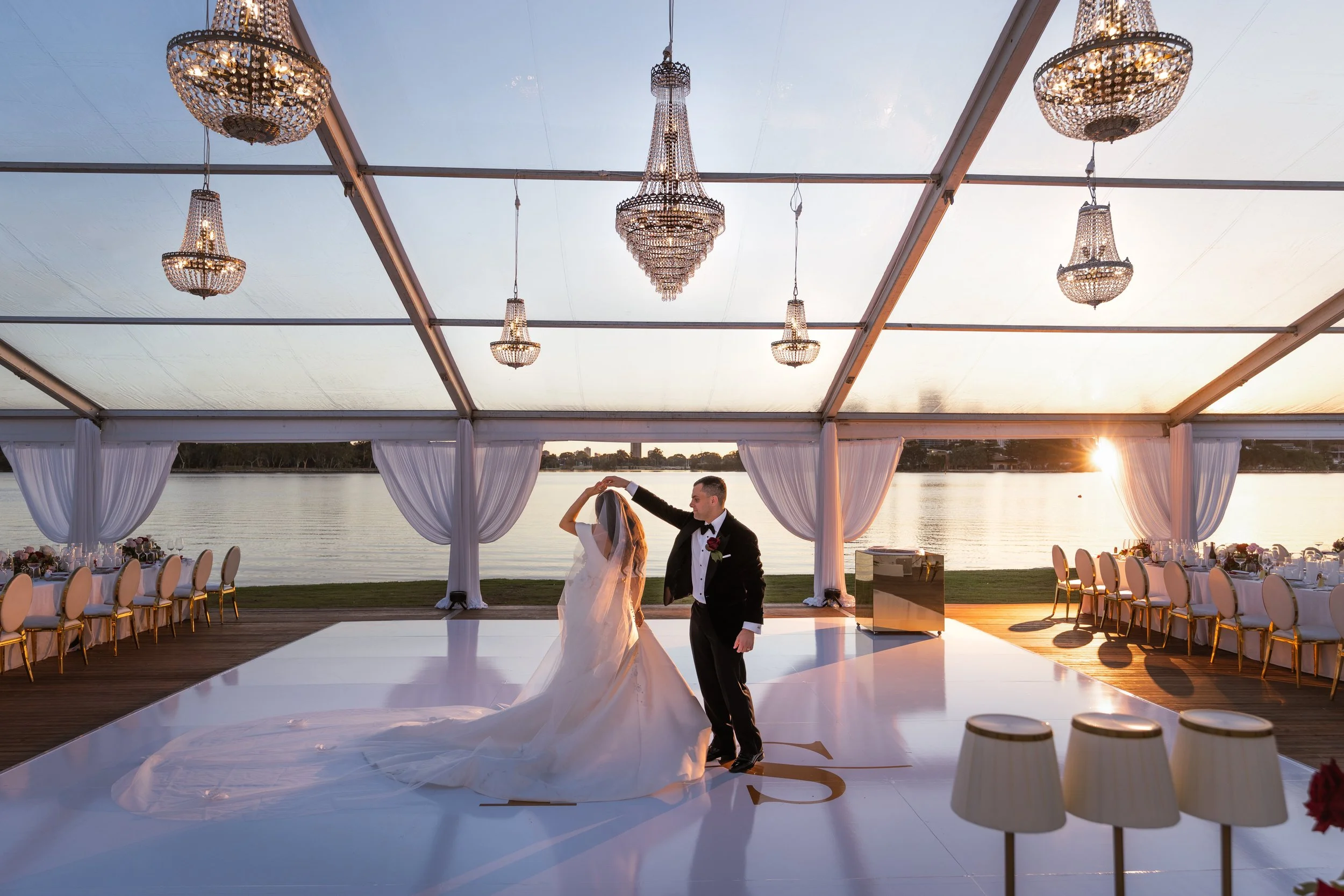 Groom spinning bride on a monogrammed white dance floor inside a crystal-chandelier marquee with a golden river sunset behind
