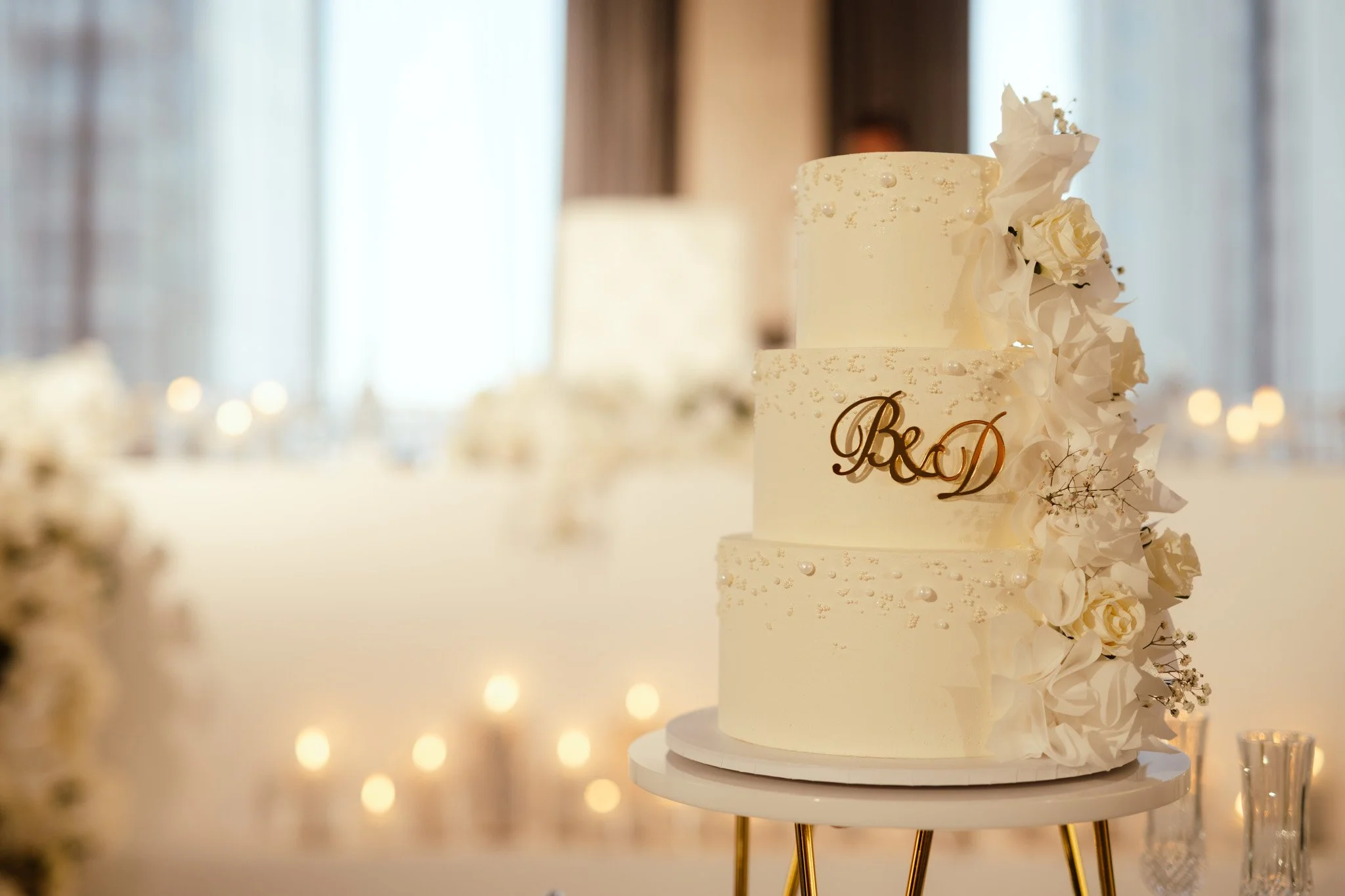 Elegant three-tier white wedding cake decorated with pearl details, white roses, and a gold B&D monogram topper, displayed in a luxurious Perth wedding reception at Crown, photographed by James Di Stefano