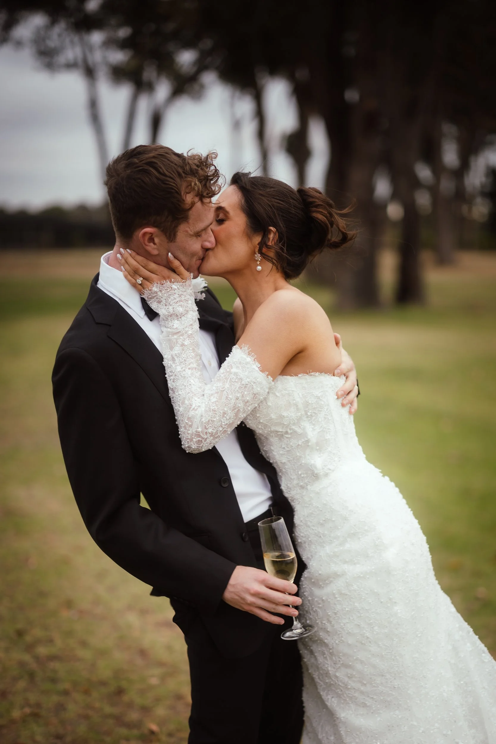 Bride in a beaded off-shoulder gown and lace gloves pulls her groom in for a kiss while holding a champagne flute, tall