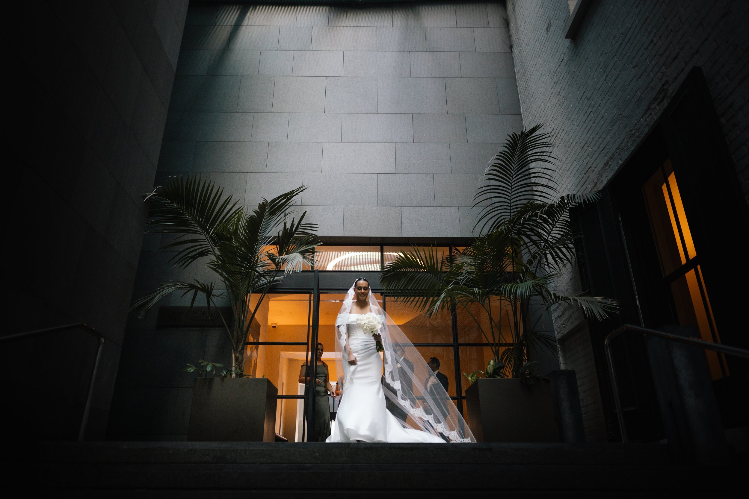 A beaming bride in a fitted off-shoulder gown and flowing cathedral veil walks down grand stone steps framed by tall palms.