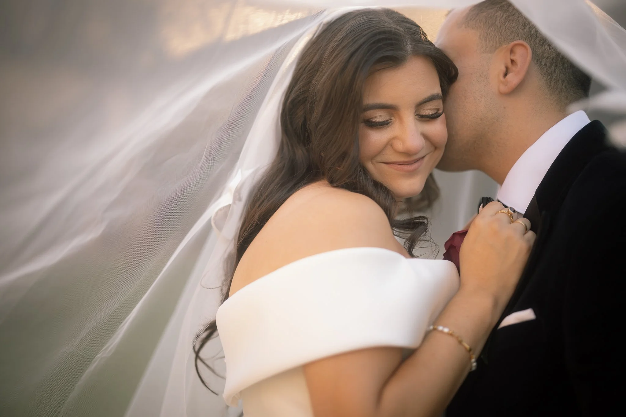 A glowing bride smiles softly as her groom kisses her cheek, both draped beneath a billowing tulle veil.
