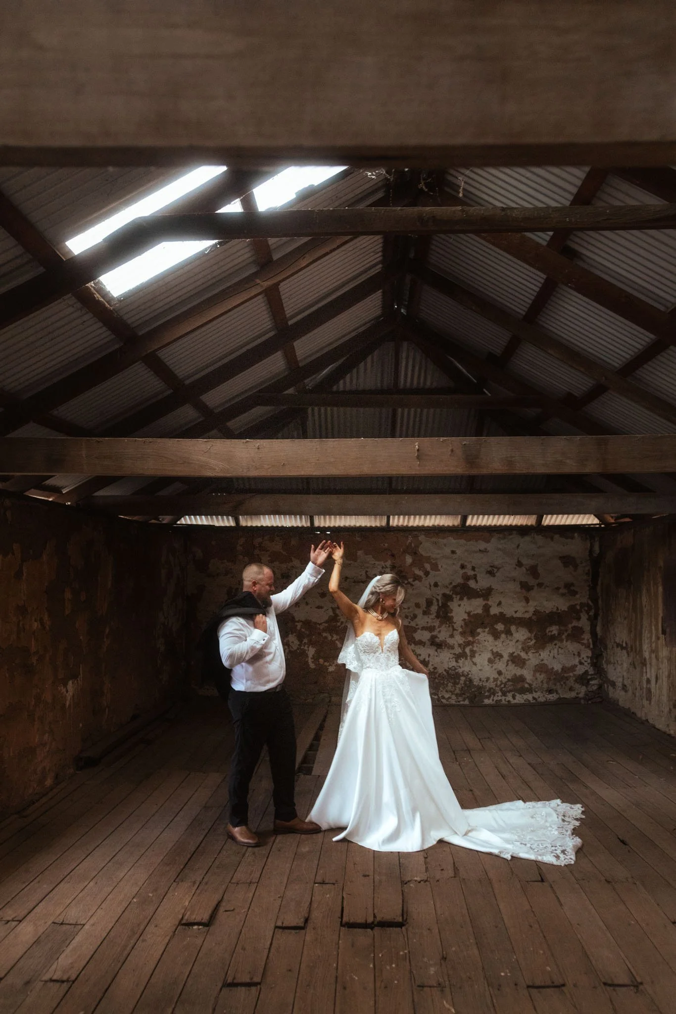 Inside the rustic stone barn at Buckland Estate, a groom twirls his bride on the vintage timber floorboards under an exposed beam roof, captured in a moody, editorial style by a luxury Perth wedding photographer