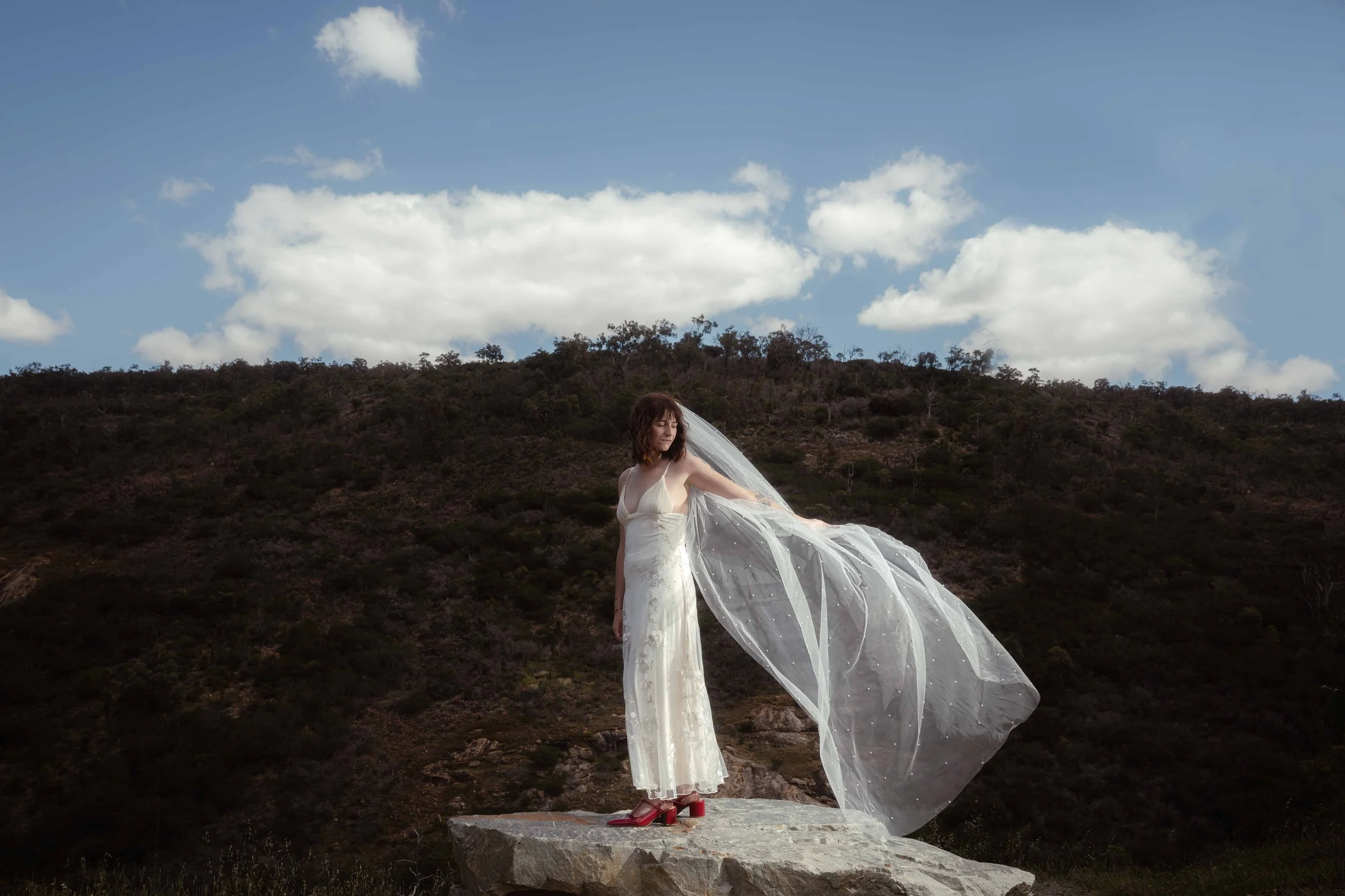 A bride in a floral slip dress with a pearl-dotted veil sweeping dramatically in the breeze atop a granite boulder