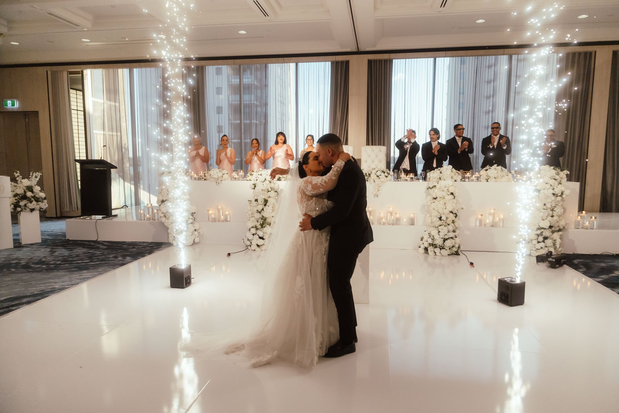Bride and groom sharing a passionate kiss during their first dance on a white reflective dance floor at Crown Perth, surrounded by twin cold sparkler fountains and their cheering bridal party, photographed by James Di Stefano