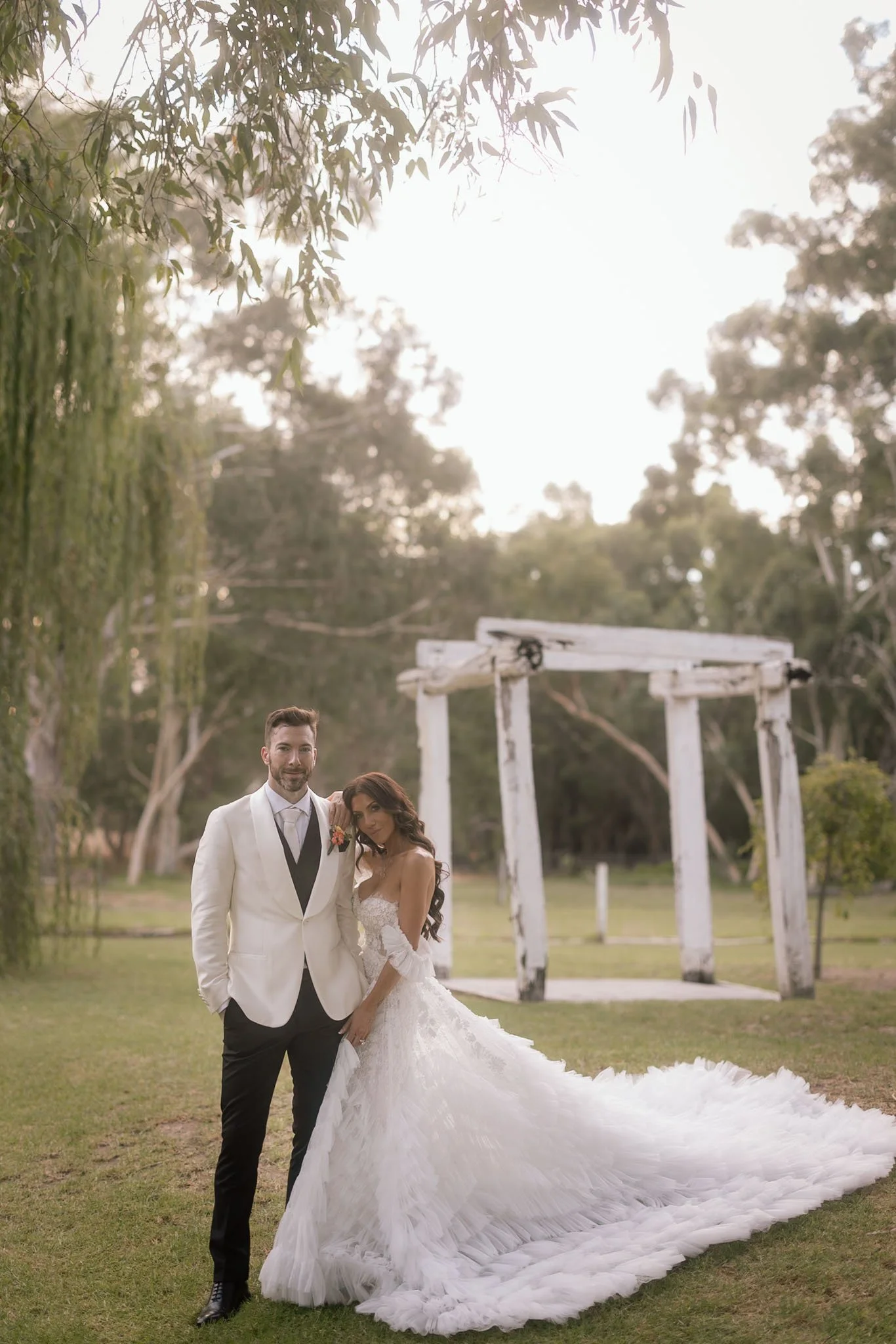 Bride and groom posing together in front of a rustic white timber arch at The Mallard Duck Swan Valley at golden hour, surrounded by willow trees, photographed by James Di Stefano