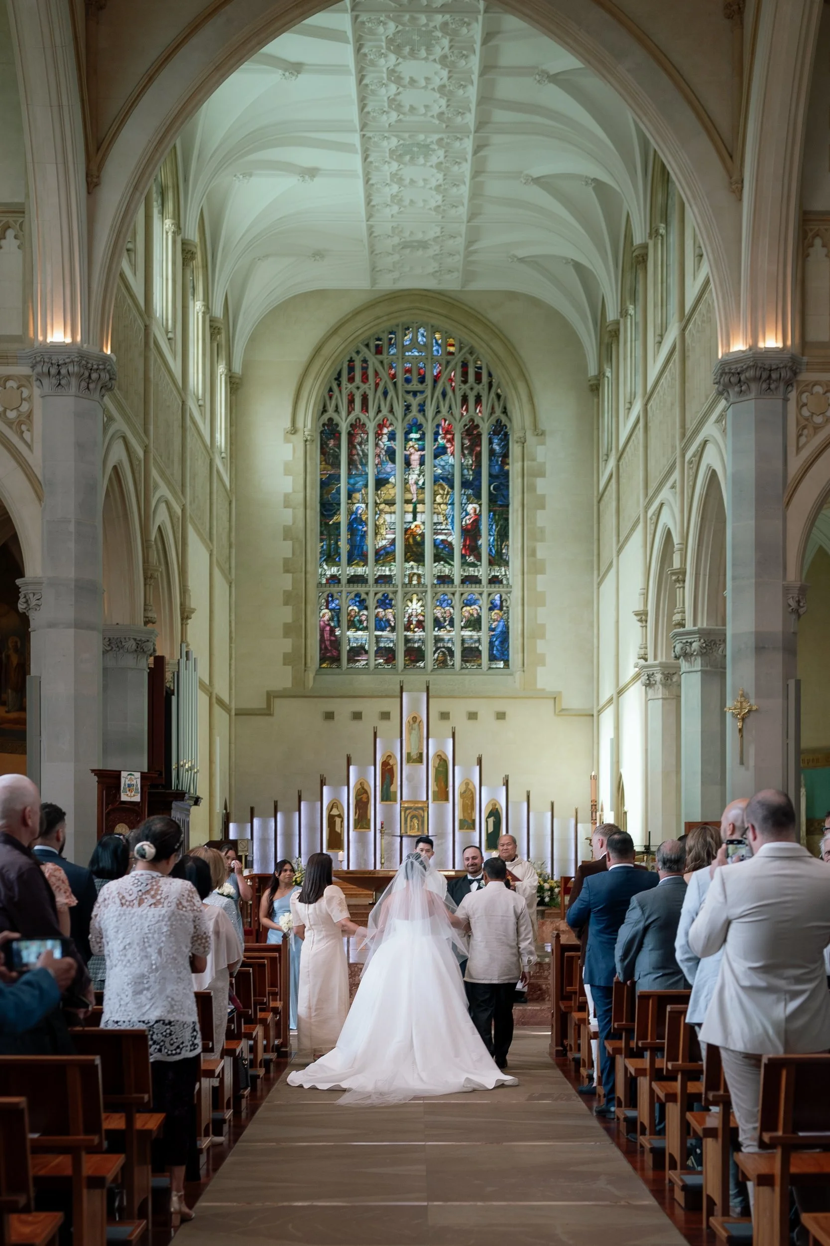 A bride in a long veil walks down the aisle of a grand Gothic cathedral toward her groom, a stunning stained-glass window