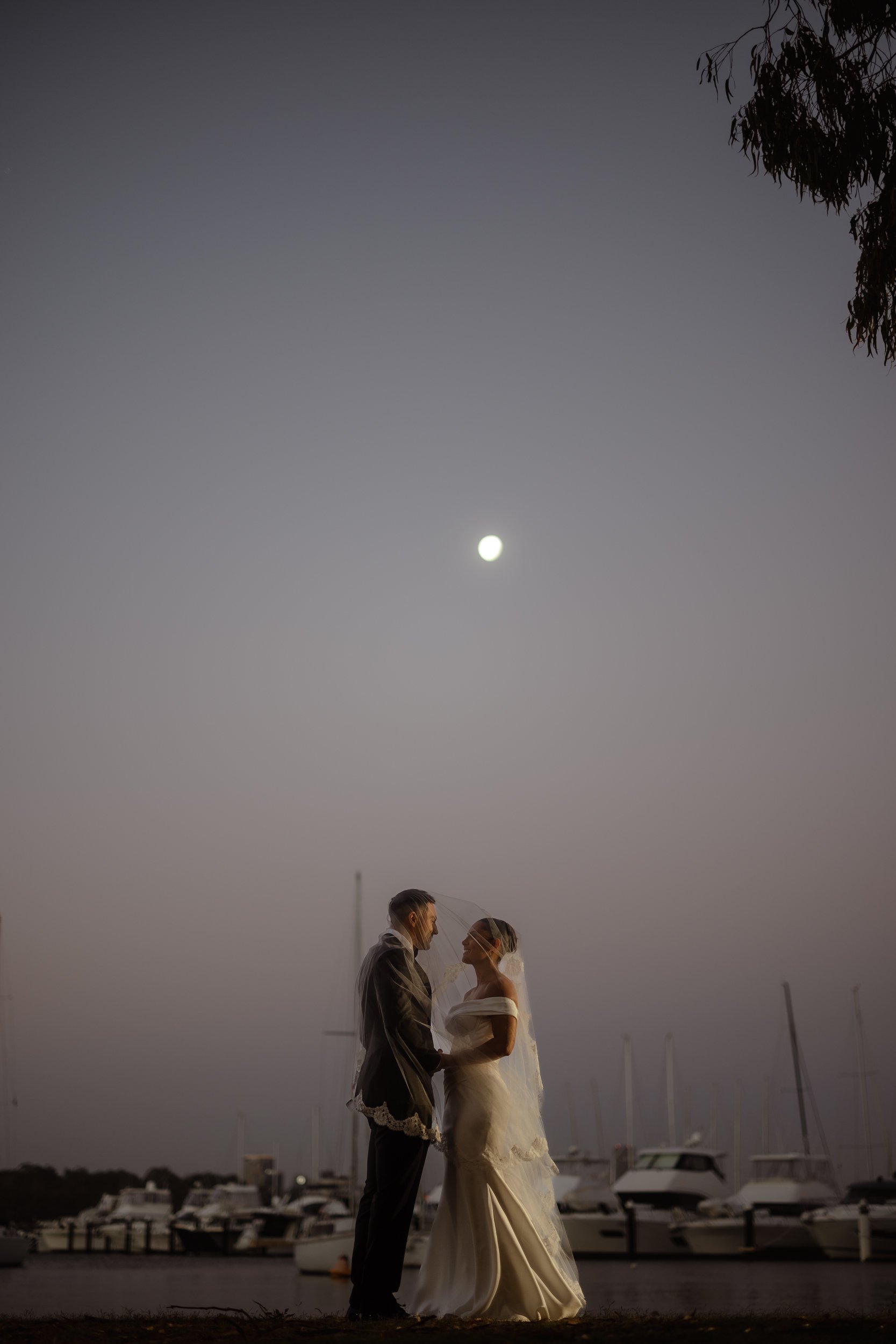 Bride in an off-shoulder gown and long veil gazes at her groom beneath a full moon, moody marina boats visible behind them.