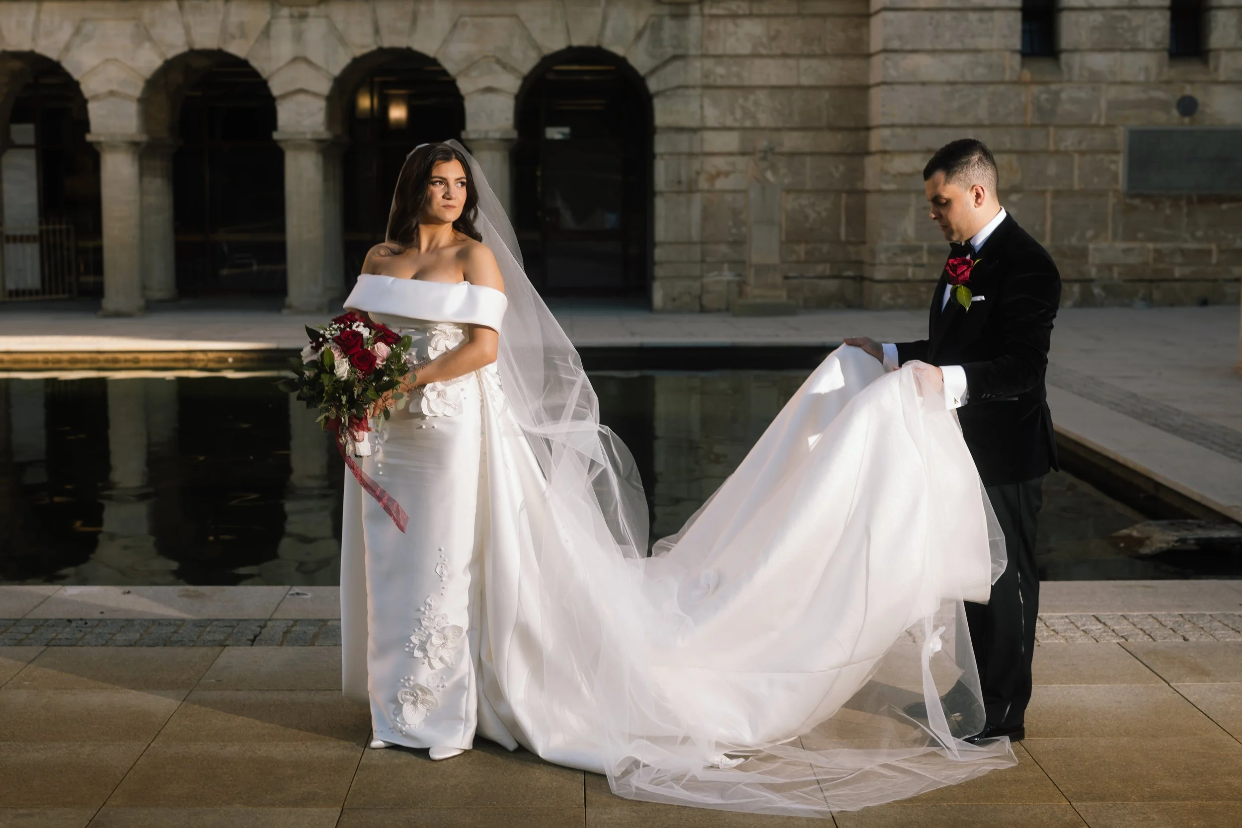 Groom lifts the sweeping cathedral train of his bride's off-shoulder gown beside a sandstone colonnade