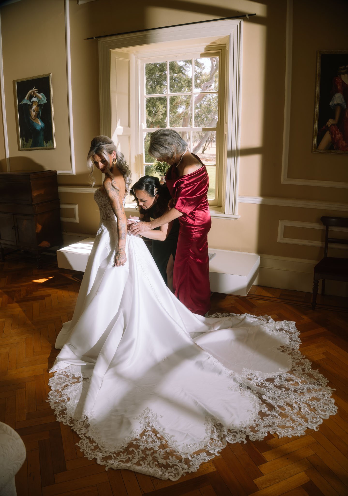 A luxury bride in an ornate lace wedding gown with a dramatic long train being assisted by her bridal party inside a sun-drenched heritage room with vintage parquet flooring at Buckland Estate.