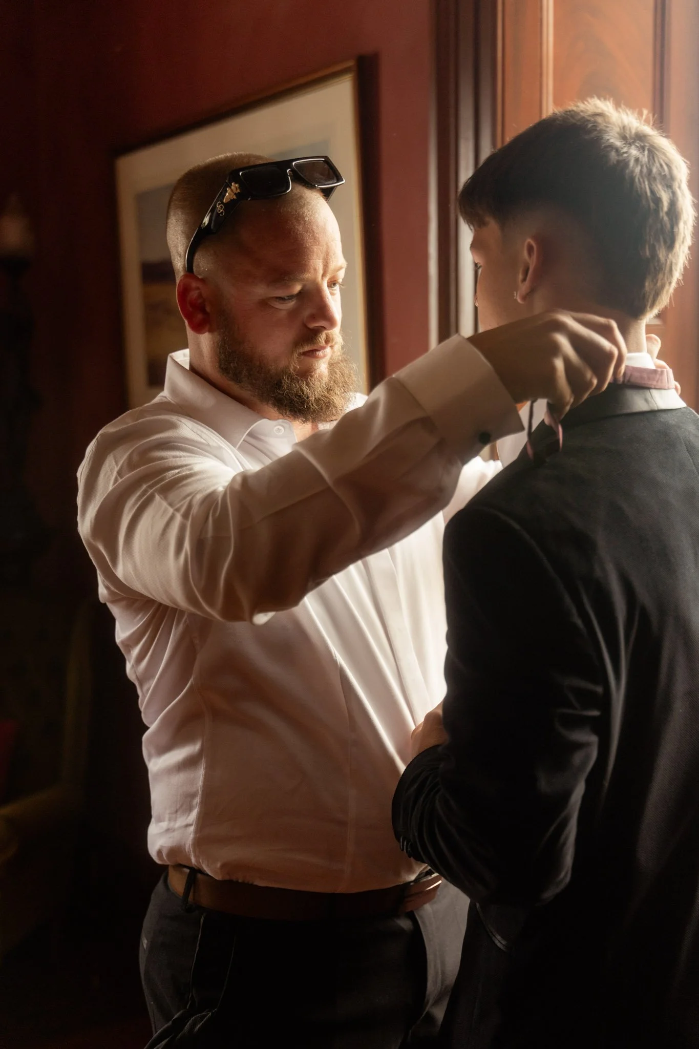 Candid morning preparation moment of a groom adjusting a groomsman's bowtie inside the vintage accommodations at Buckland Estate, captured in soft natural window lighting.