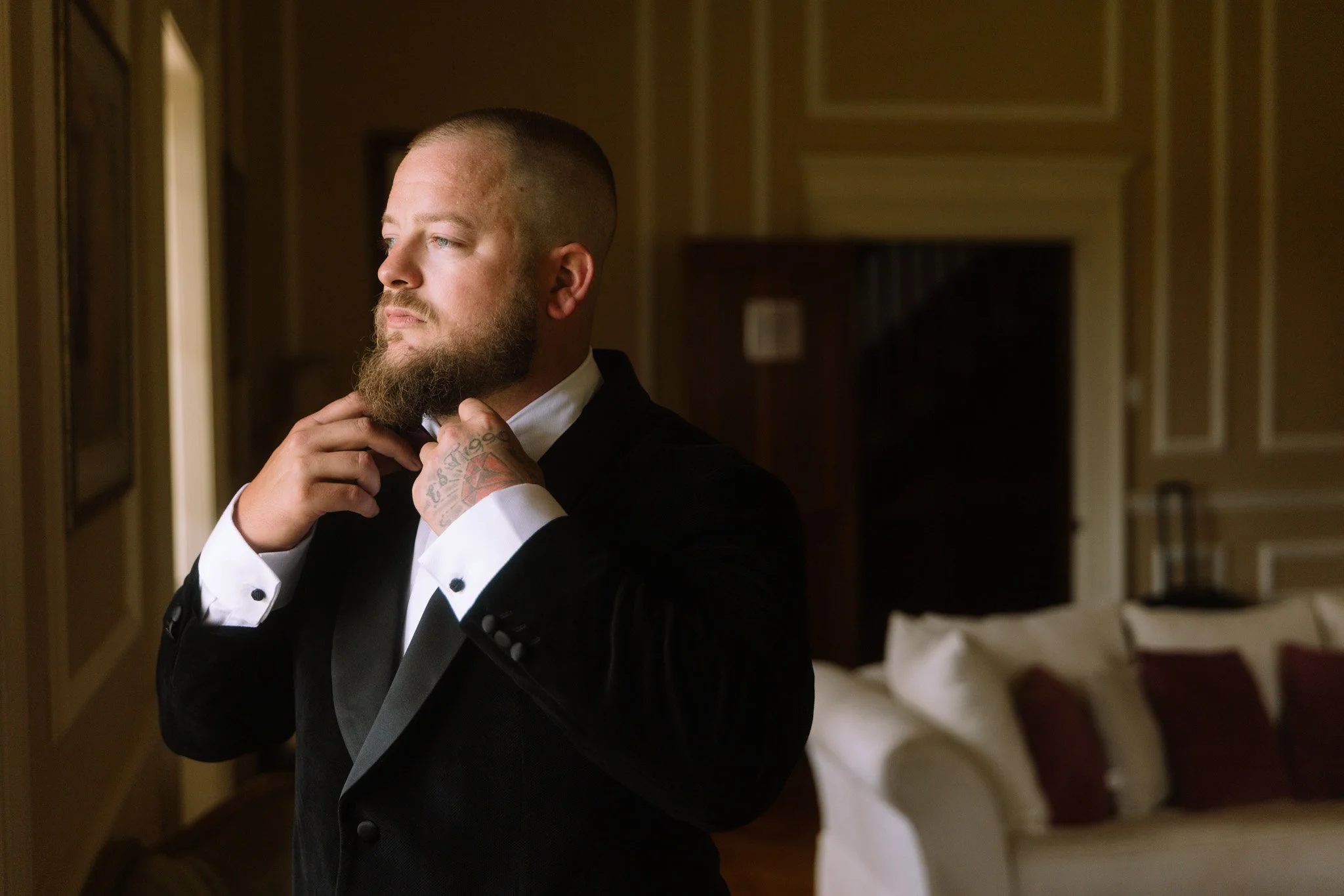 A luxury groom wearing a black velvet tuxedo looking out a grand wooden window inside Buckland Estate manor house, utilizing dramatic directional side lighting for a timeless vintage portrait.