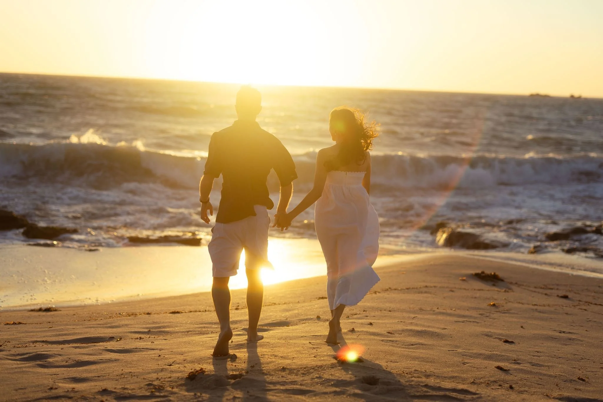 A couple walks hand-in-hand along a windswept beach at golden hour, her white dress catching the breeze during a romantic pre wedding shoot