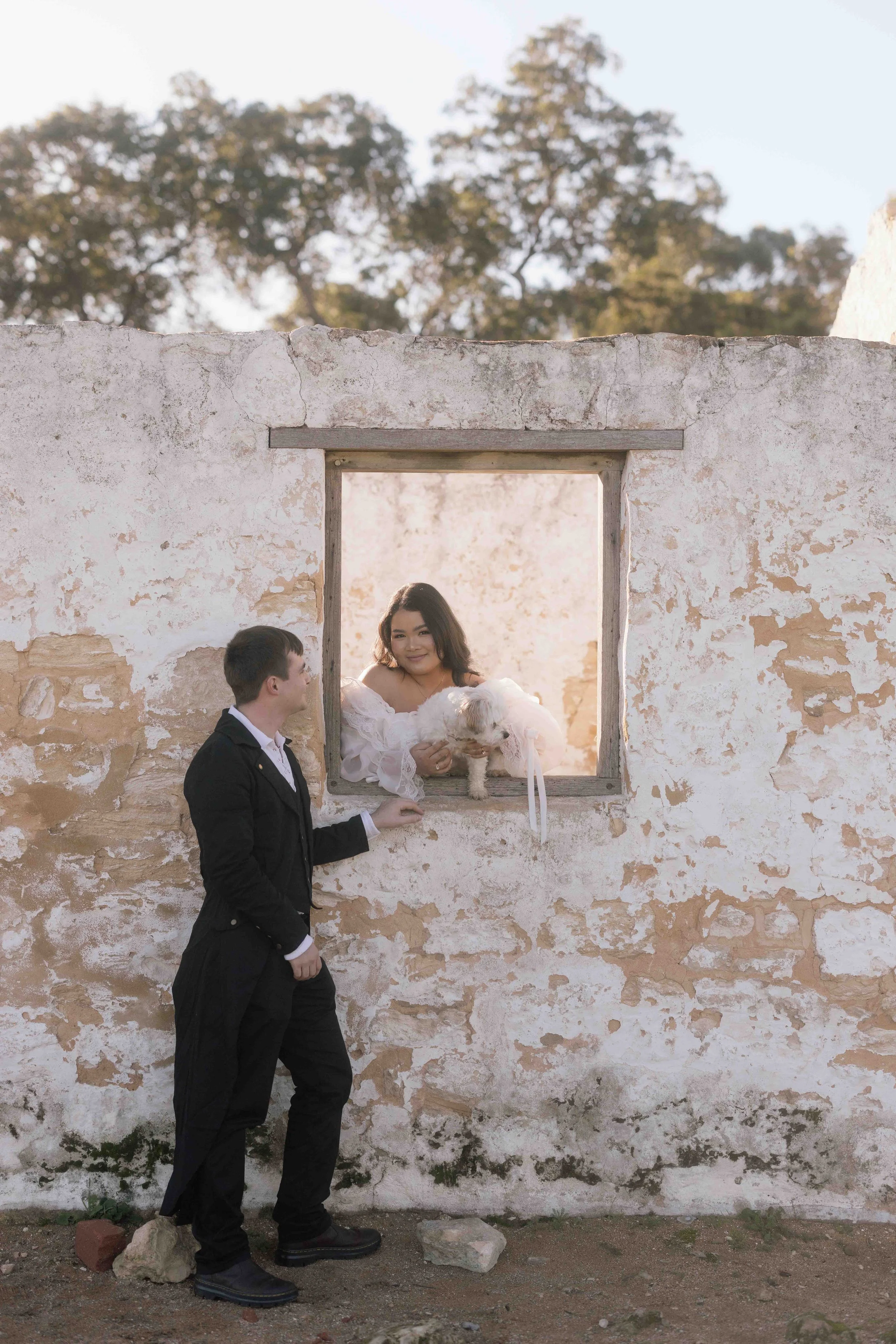 Bride cradling a small white dog leans through a weathered stone window at Perry's Paddock as her groom looks up adoringly.