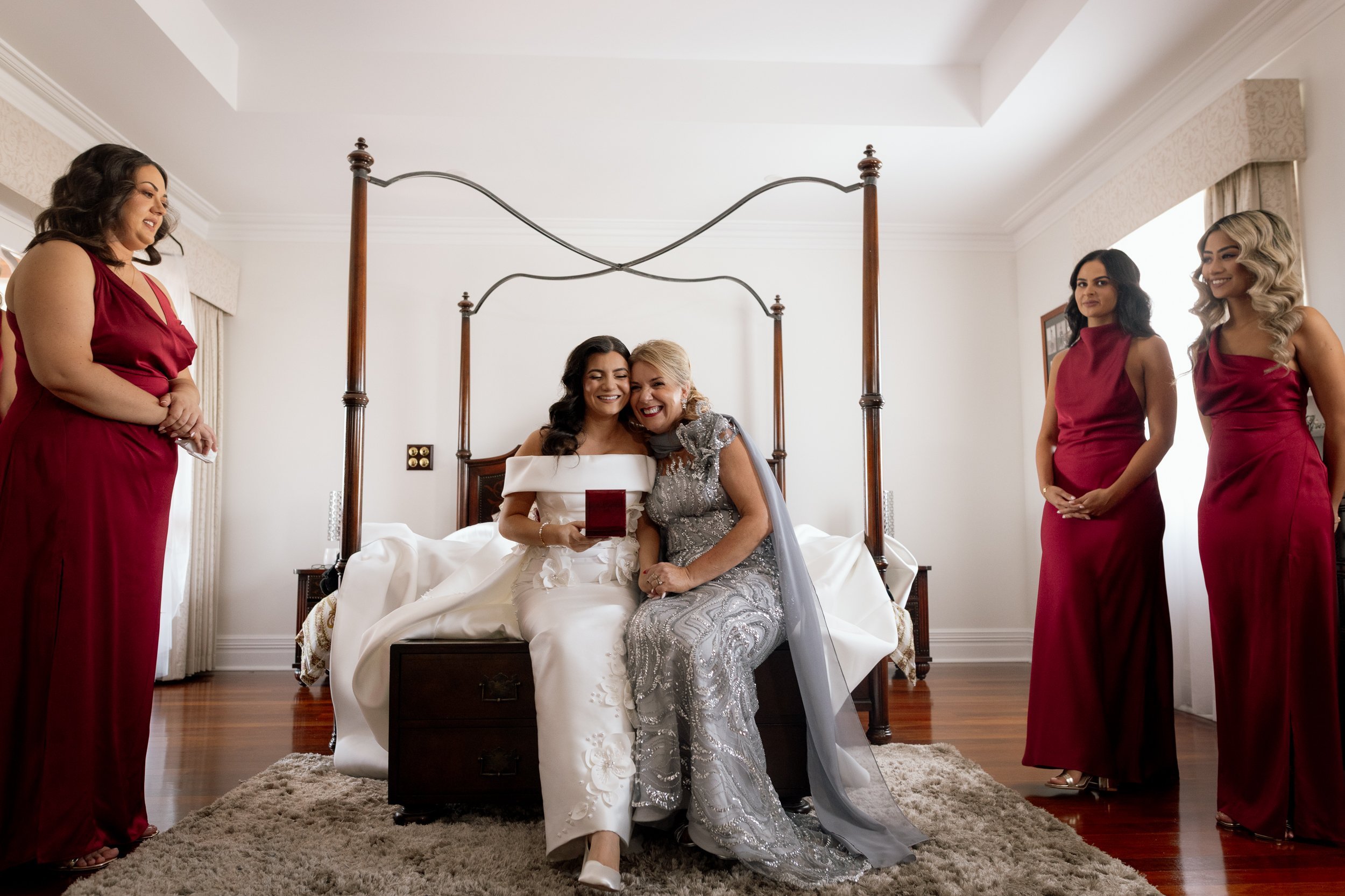 Bride in an off-shoulder white gown laughs with her mother on a four-poster bed, surrounded by bridesmaids in crimson