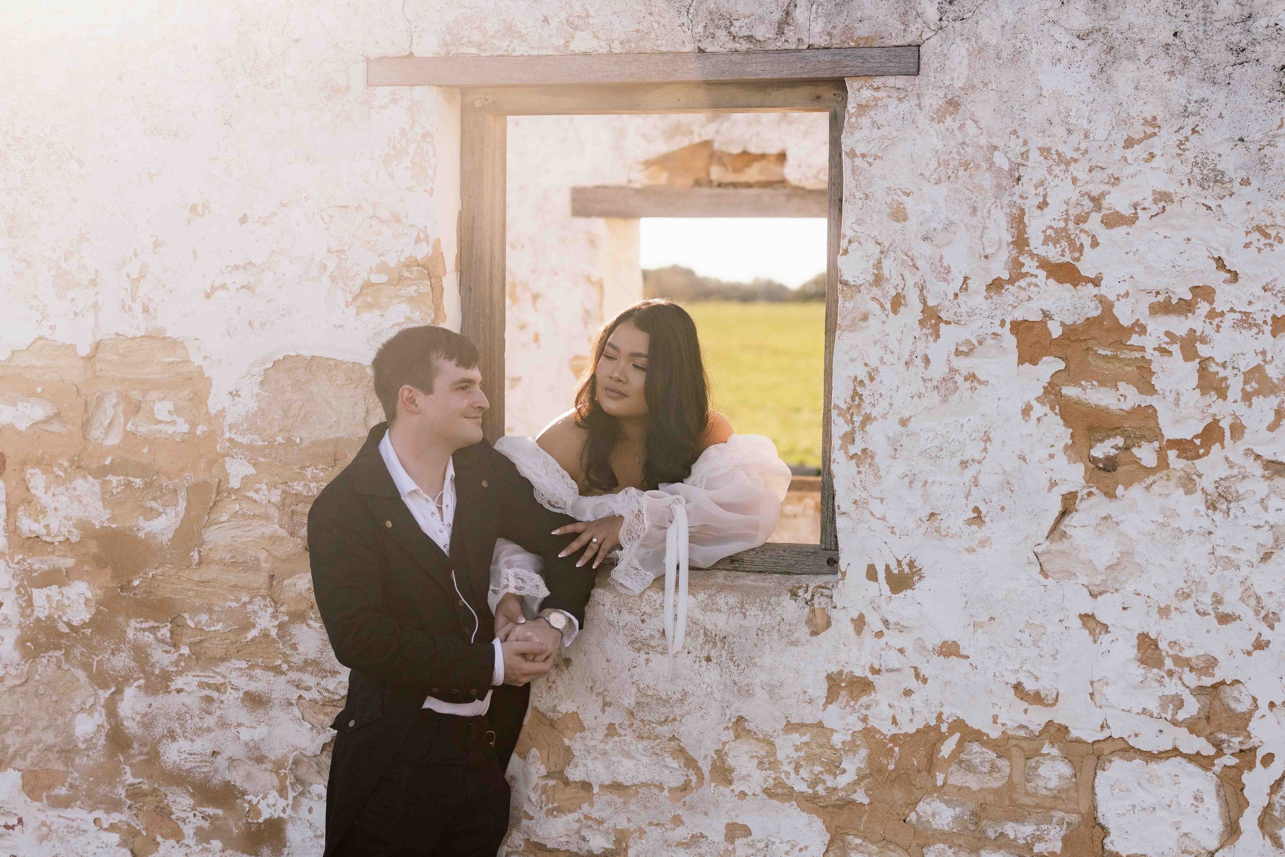 Bride leans through a timber-framed stone window clasping her groom's hands during a romantic Perry's Paddock pre-wedding shoot