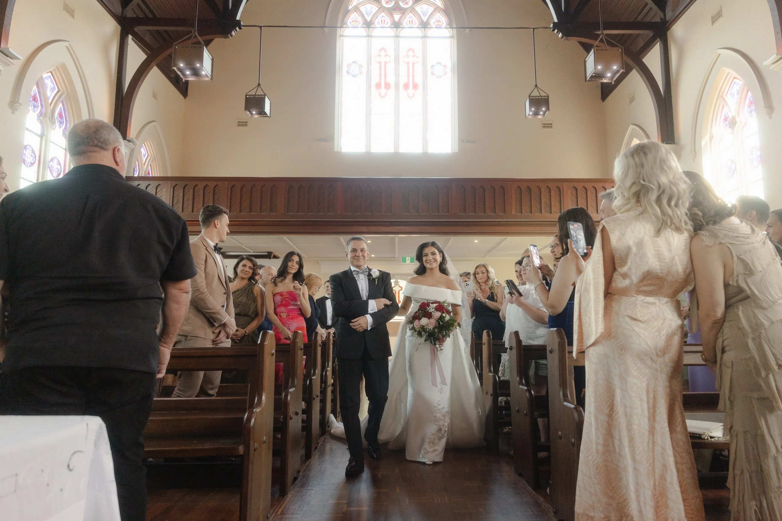 A beaming bride in an off-shoulder gown walks the aisle with her father past timber pews and glowing stained glass windows.