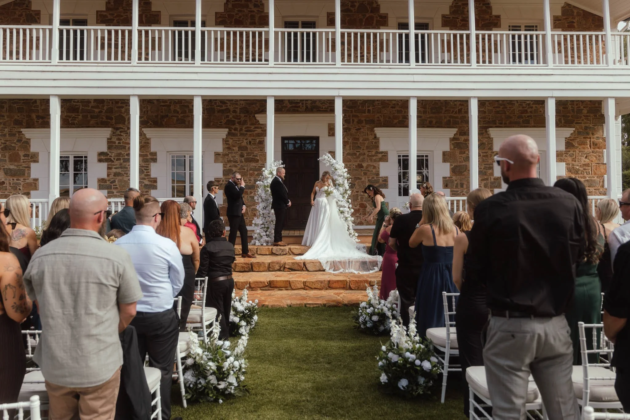 A perfectly centered wide shot capturing the intimate moment of the bride and groom exchanging vows at Buckland Estate. They are framed by two white floral arches and the symmetrical facade of the historic colonial homestead.