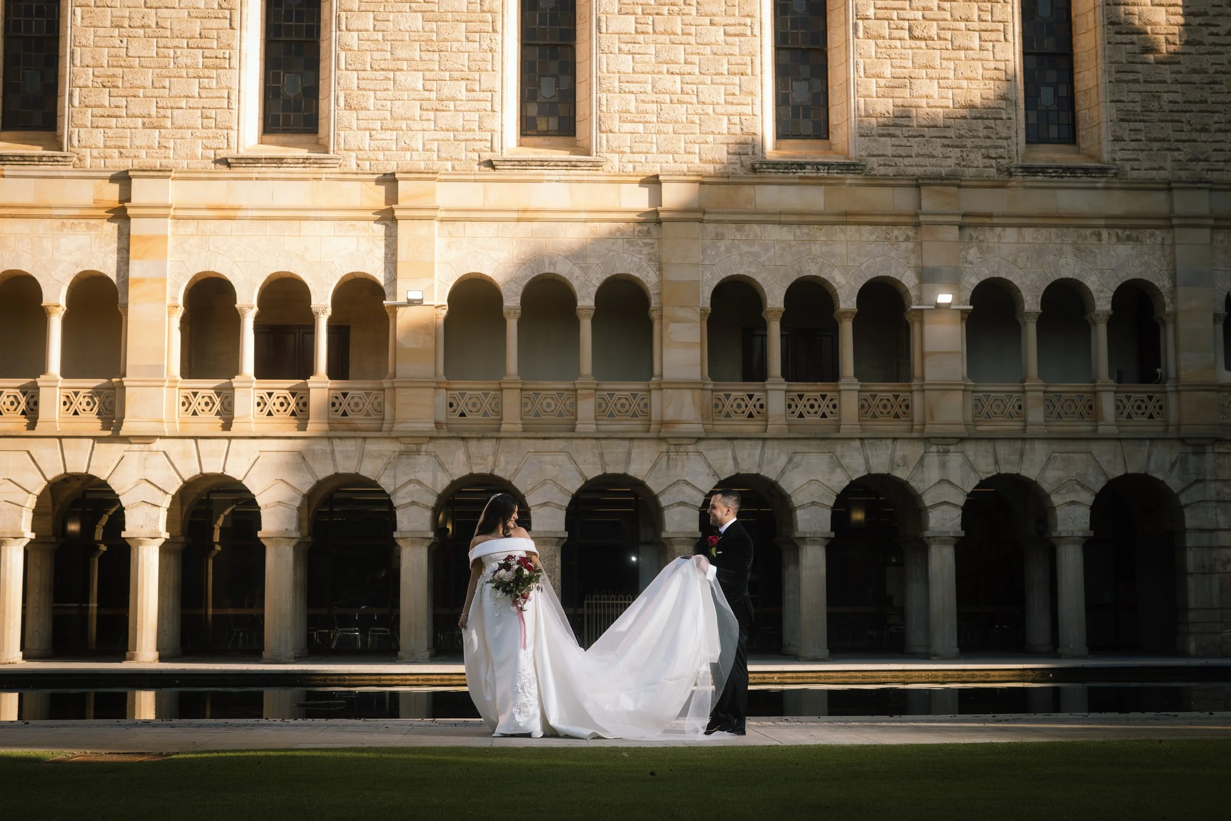 Groom lifts his bride's billowing train before the sandstone arched cloisters of UWA in this cinematic Perth wedding portrait