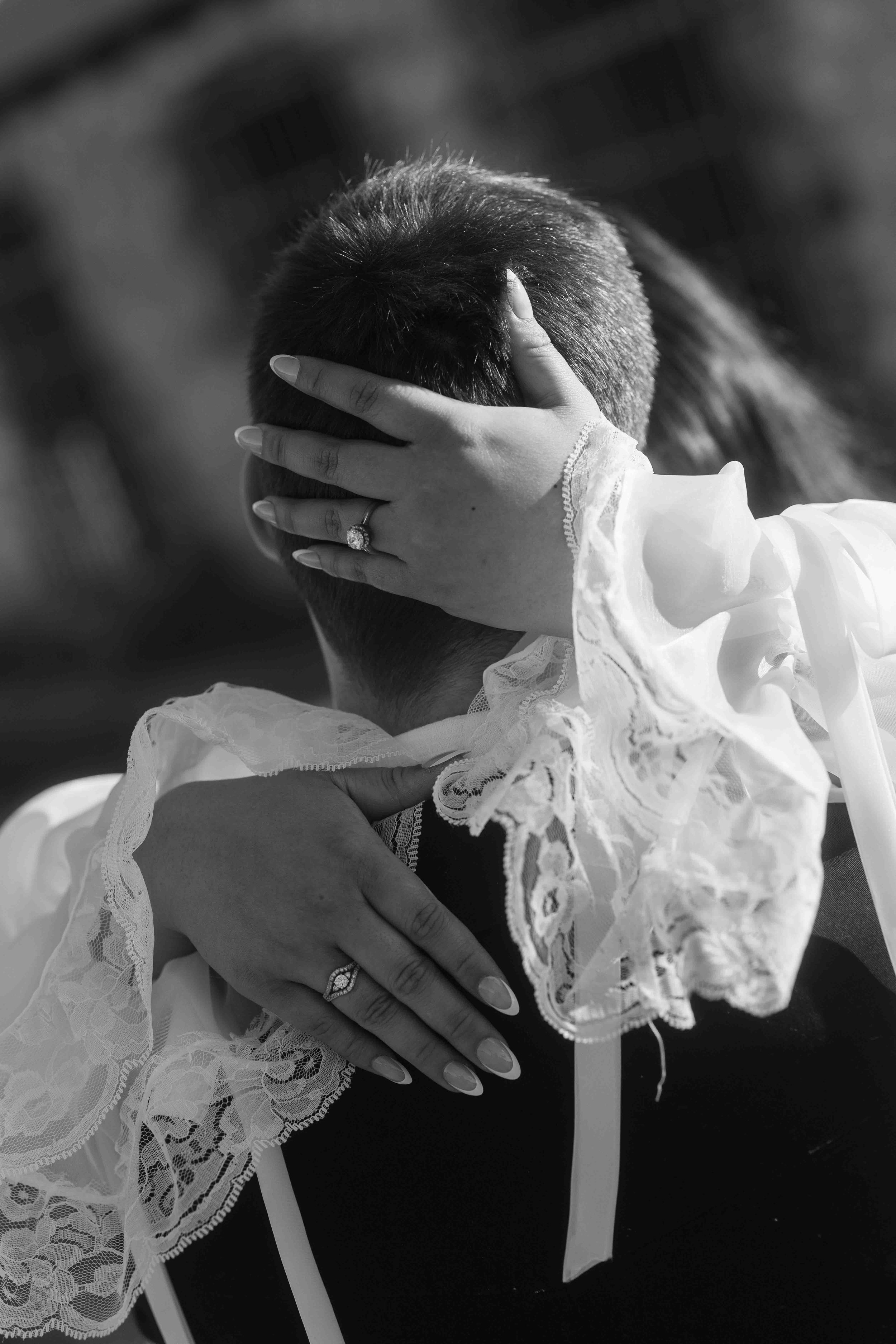 Bride's lace-cuffed hands tenderly cradle her groom, diamond engagement ring catching the light in this cinematic black and white.