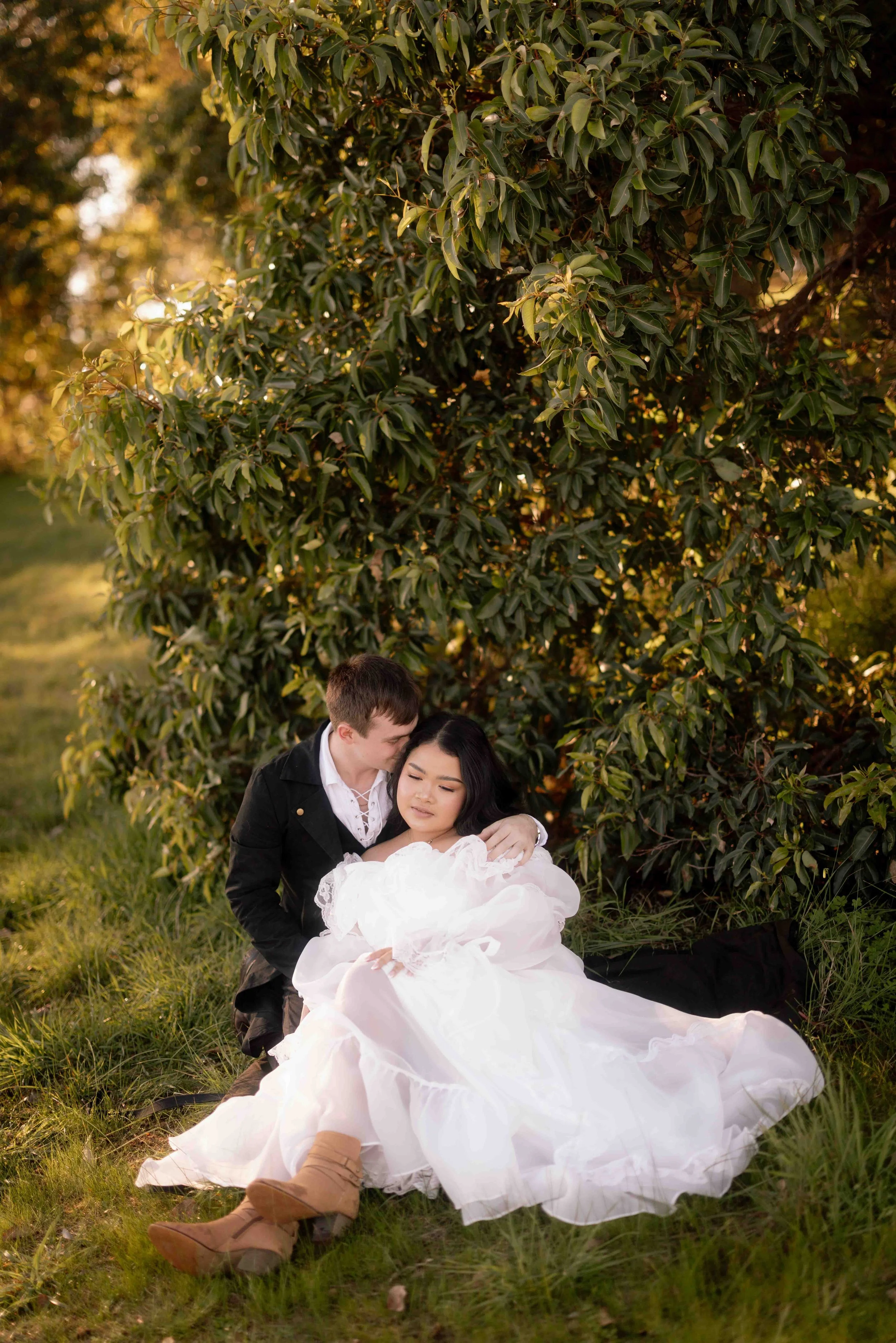 A groom gently kisses his bride's temple as she rests against him beneath a leafy tree at Perry's Paddock during golden hour.