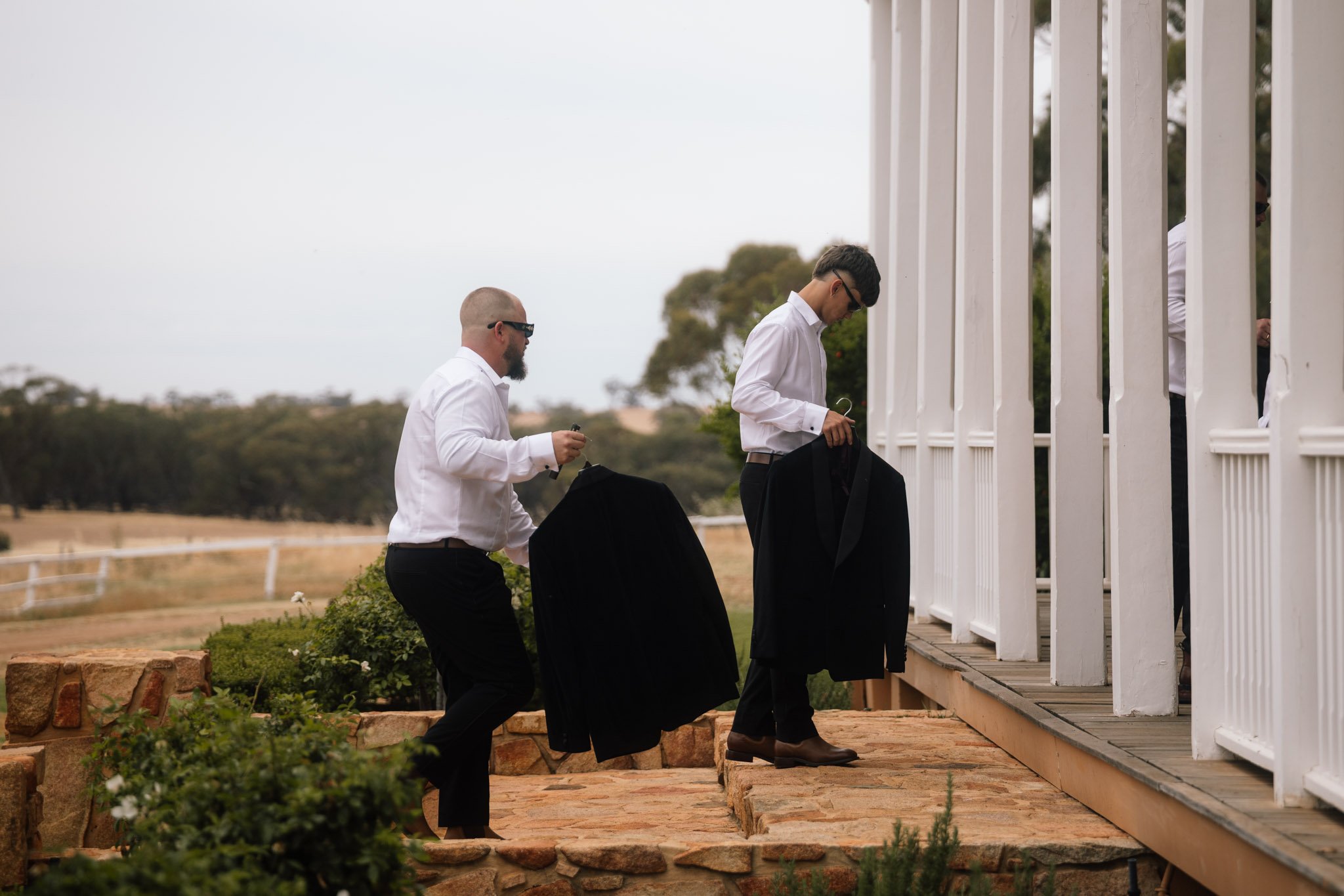 Groomsmen carrying their black tuxedo jackets up the stone steps of the historic white pillared verandah at Buckland Estate, a luxury country wedding venue in the Avon Valley, Western Australia.