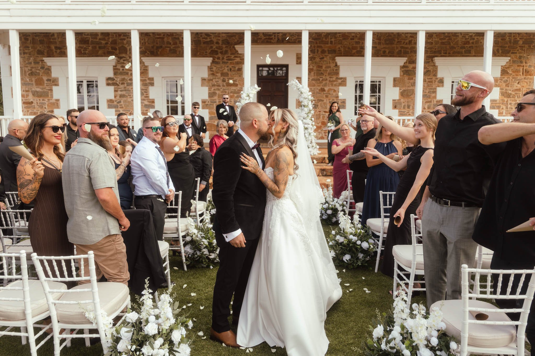 A dynamic photograph of the newly married couple walking back down the aisle at Buckland Estate, sharing a celebratory kiss while guests throw white flower petals around them on the ceremony lawn
