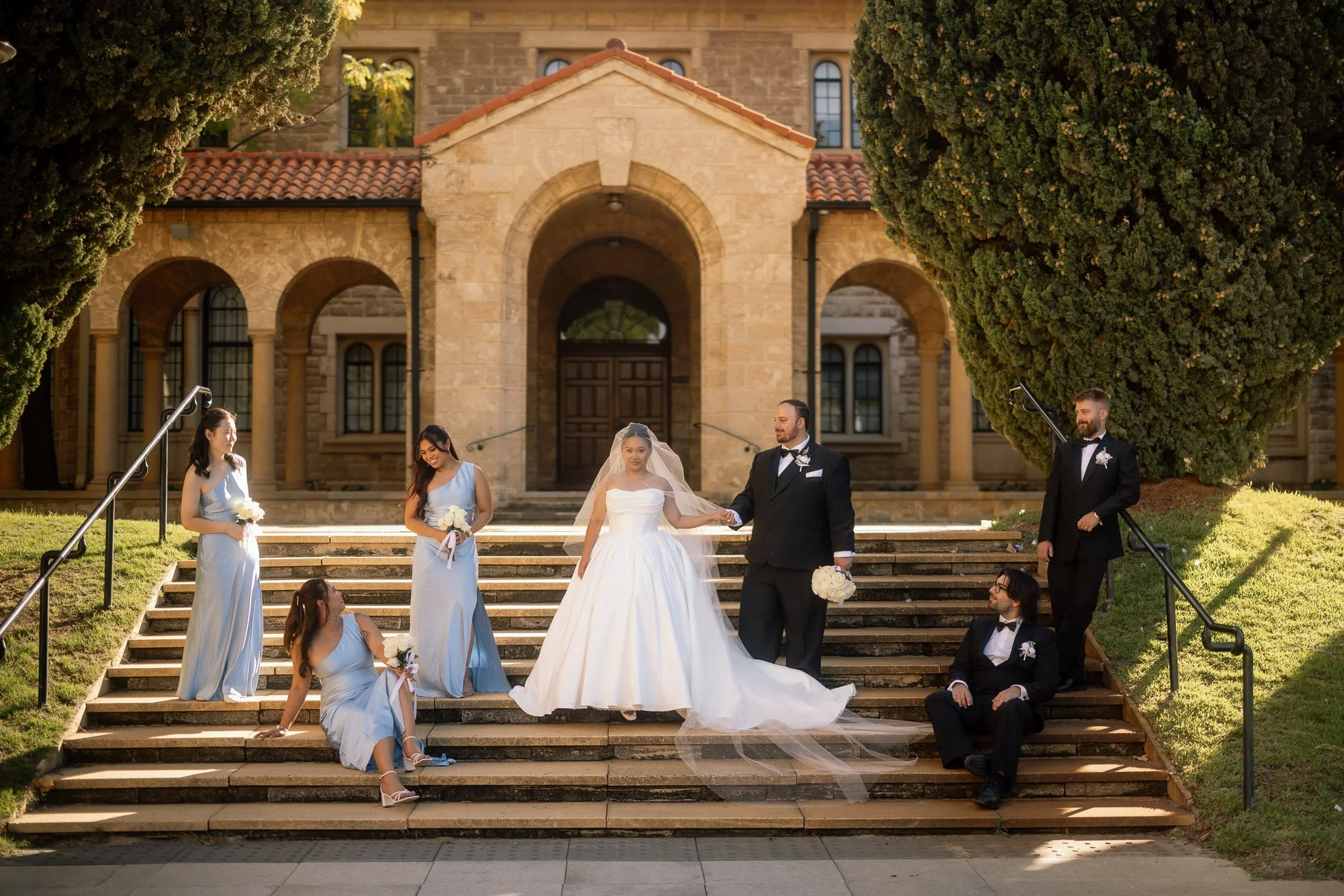 Bride and groom walk down sunlit sandstone church steps surrounded by bridesmaids in pale blue gowns and tuxedoed groomsmen.
