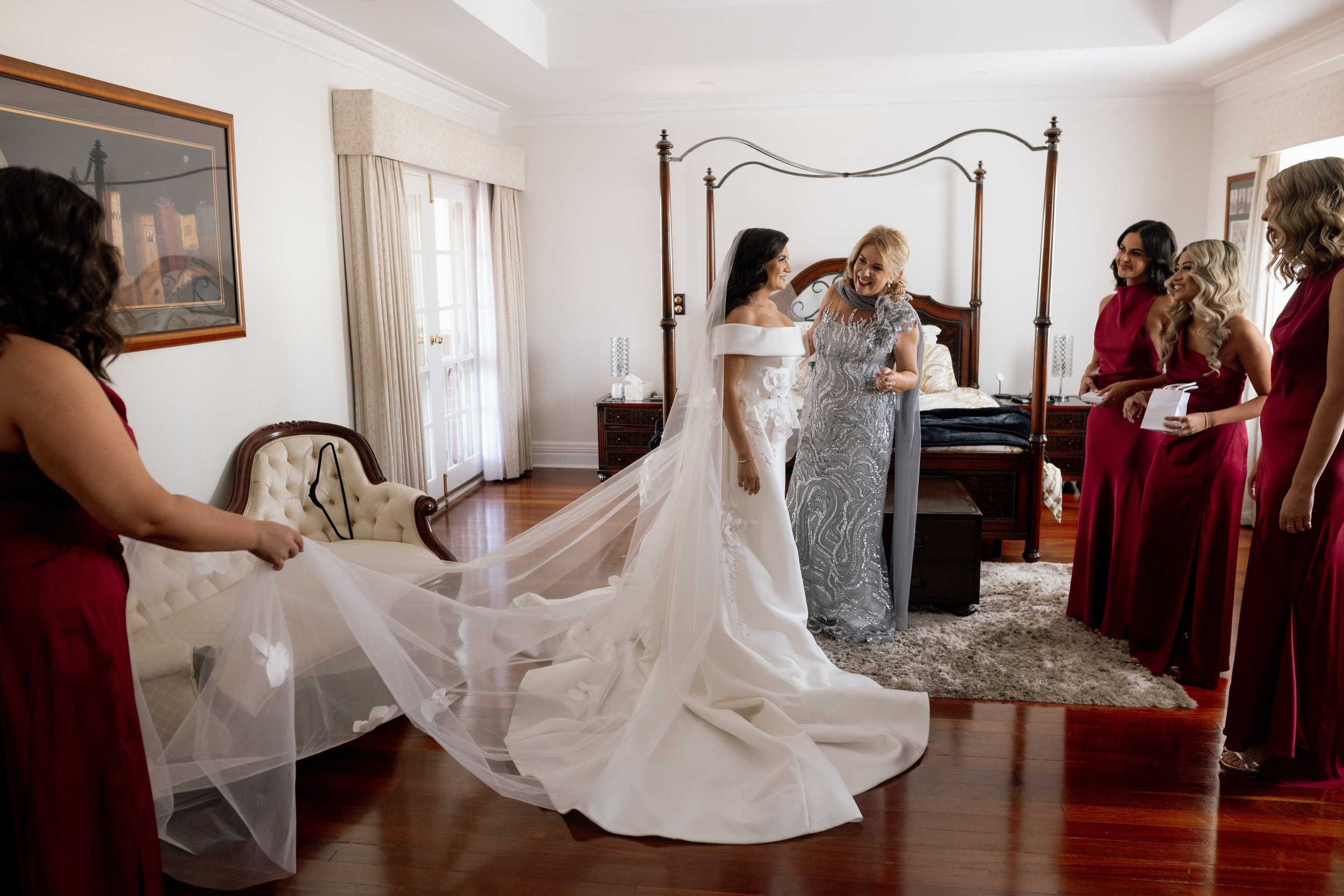 Bride in an off-shoulder gown shares a joyful moment as her bridesmaids in red carefully spread her cathedral veil during bridal preparations