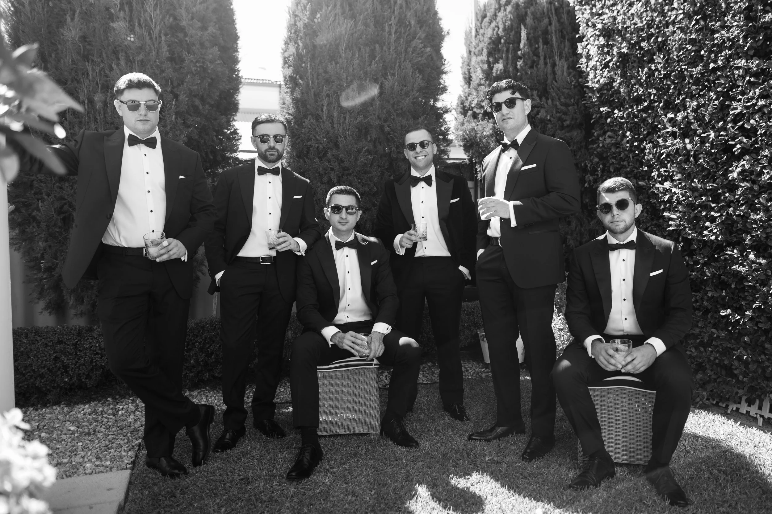 Six groomsmen in black tuxedos and sunglasses strike a cool editorial pose in a sunlit garden, captured in cinematic black and white
