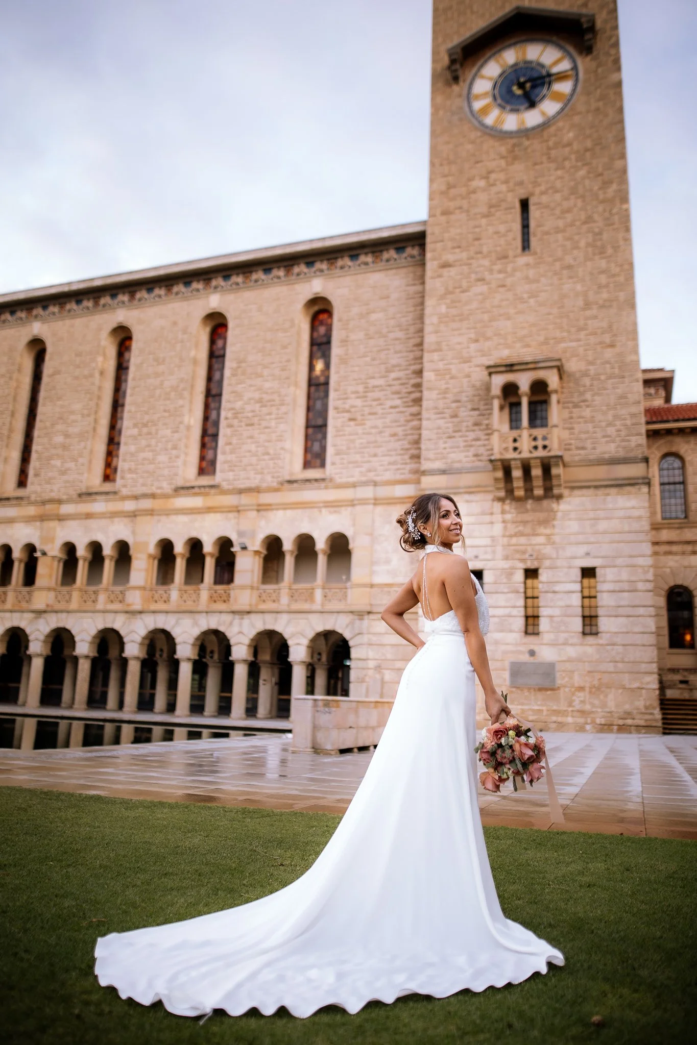 Bride in a backless halter gown and cathedral train glances over her shoulder at the UWA clock tower.