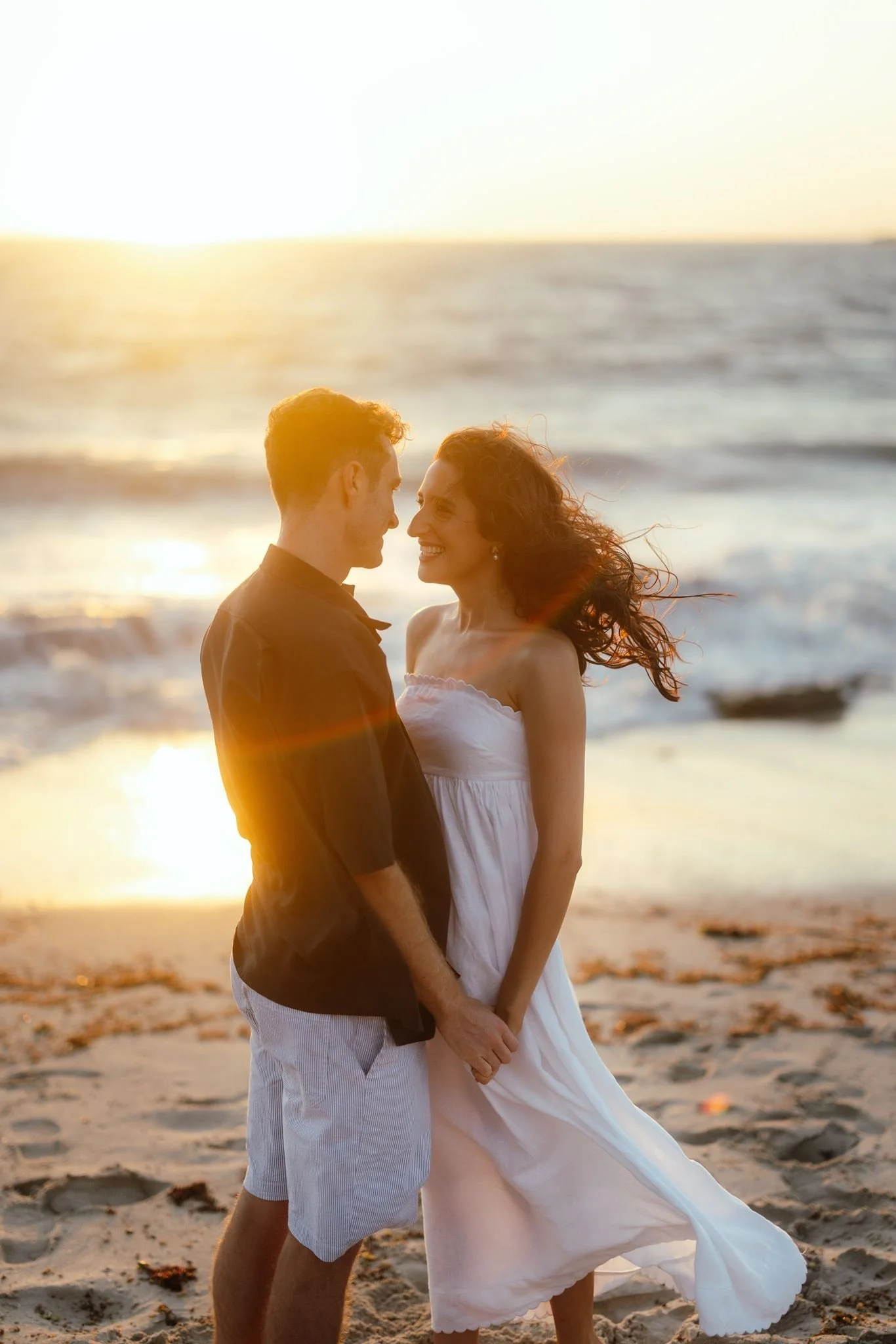 A couple share a tender moment on a windswept beach at golden hour, her white strapless dress catching the breeze during their shoot