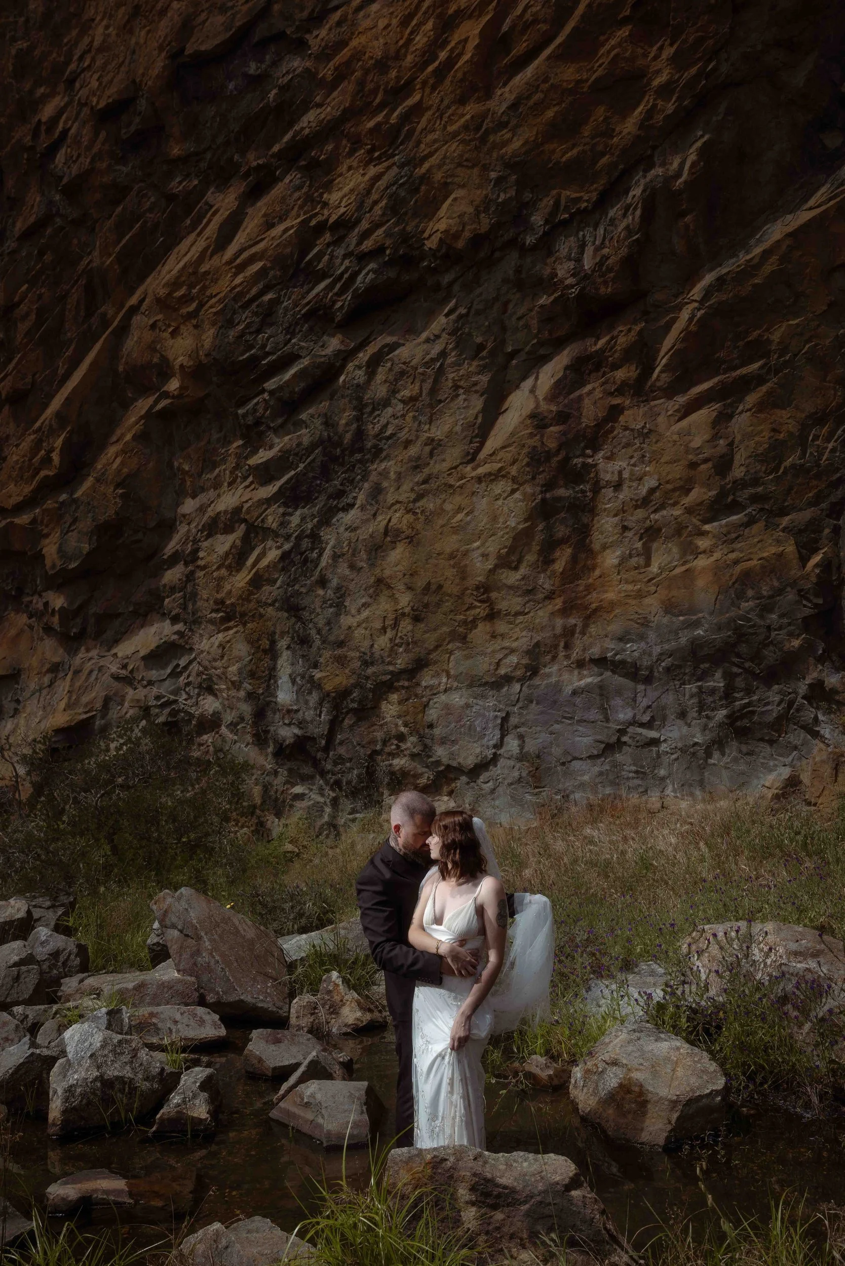 A bride in a slinky lace gown and veil embraces her groom among boulders beside a stream, framed by a dramatic sandstone