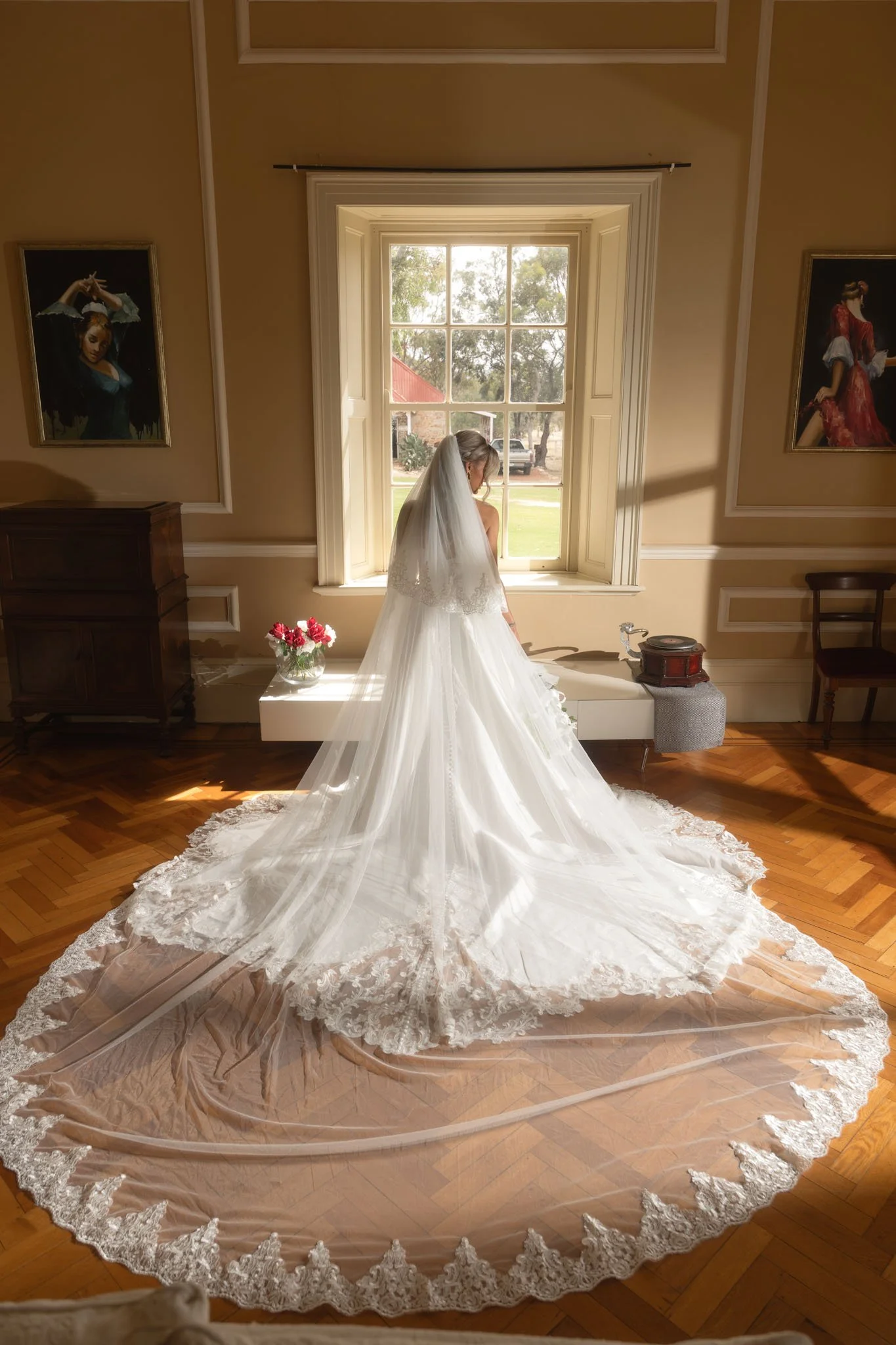 Breathtaking editorial portrait of a bride standing at a large sash window, her massive lace-trimmed wedding veil perfectly fanned out across the historic parquet floor in a pool of golden light at Buckland Estate.
