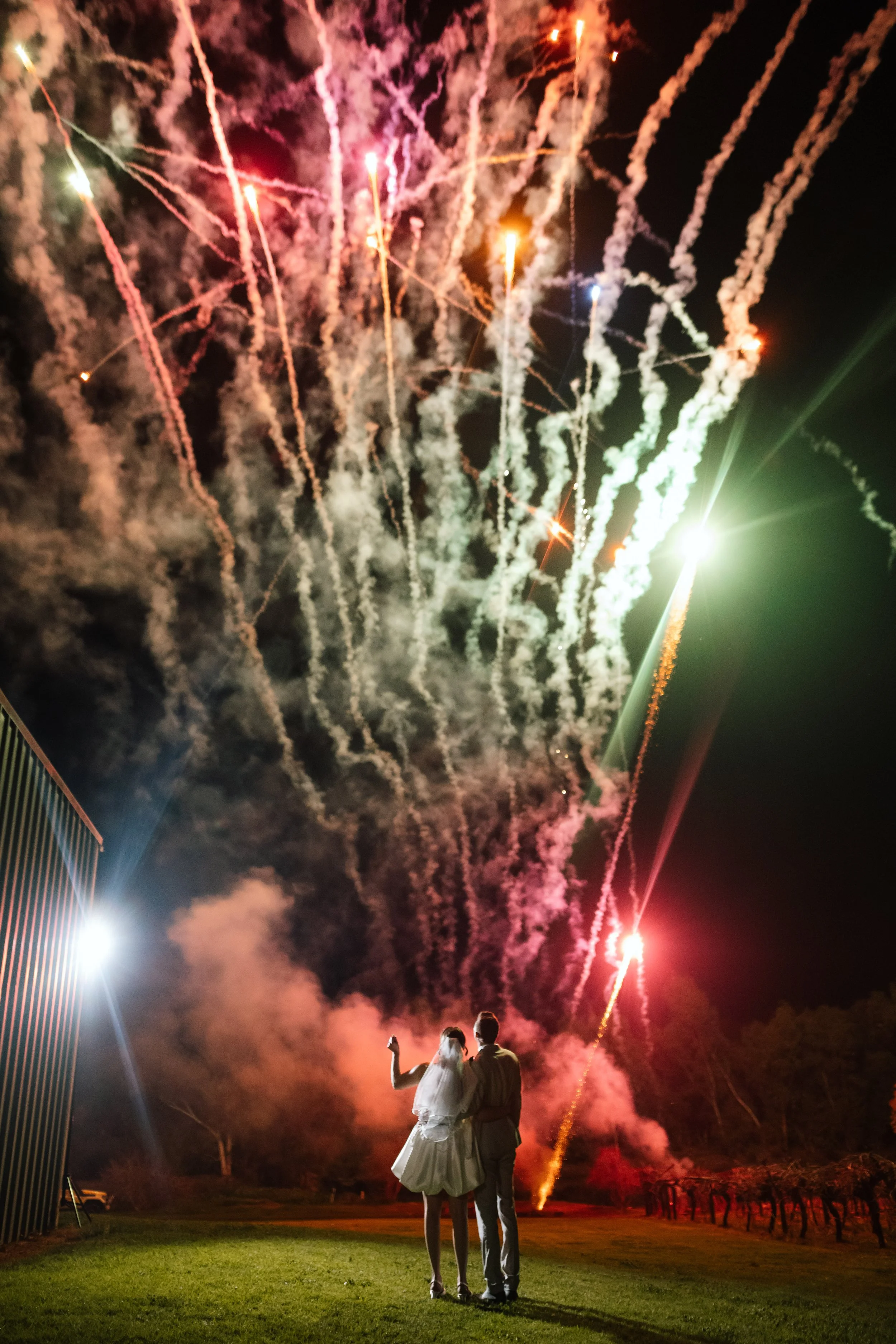 A bride and groom stand together as vivid red wedding fireworks spiral into the night sky above them, bathing the scene