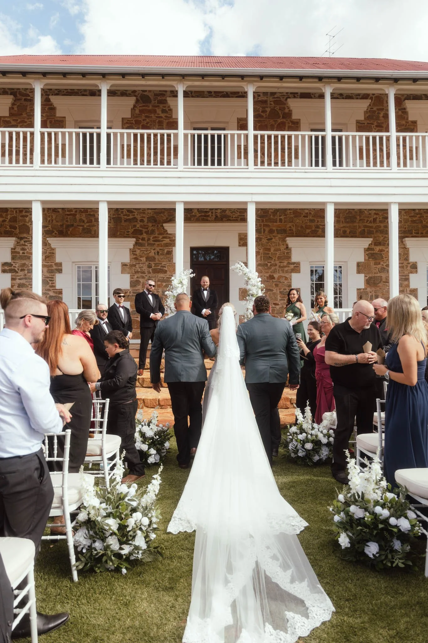 A rear-view photograph capturing the bride, with her extensive lace veil flowing behind her, walking down the grassy aisle between seated guests at Buckland Estate toward the groom and the historic homestead.