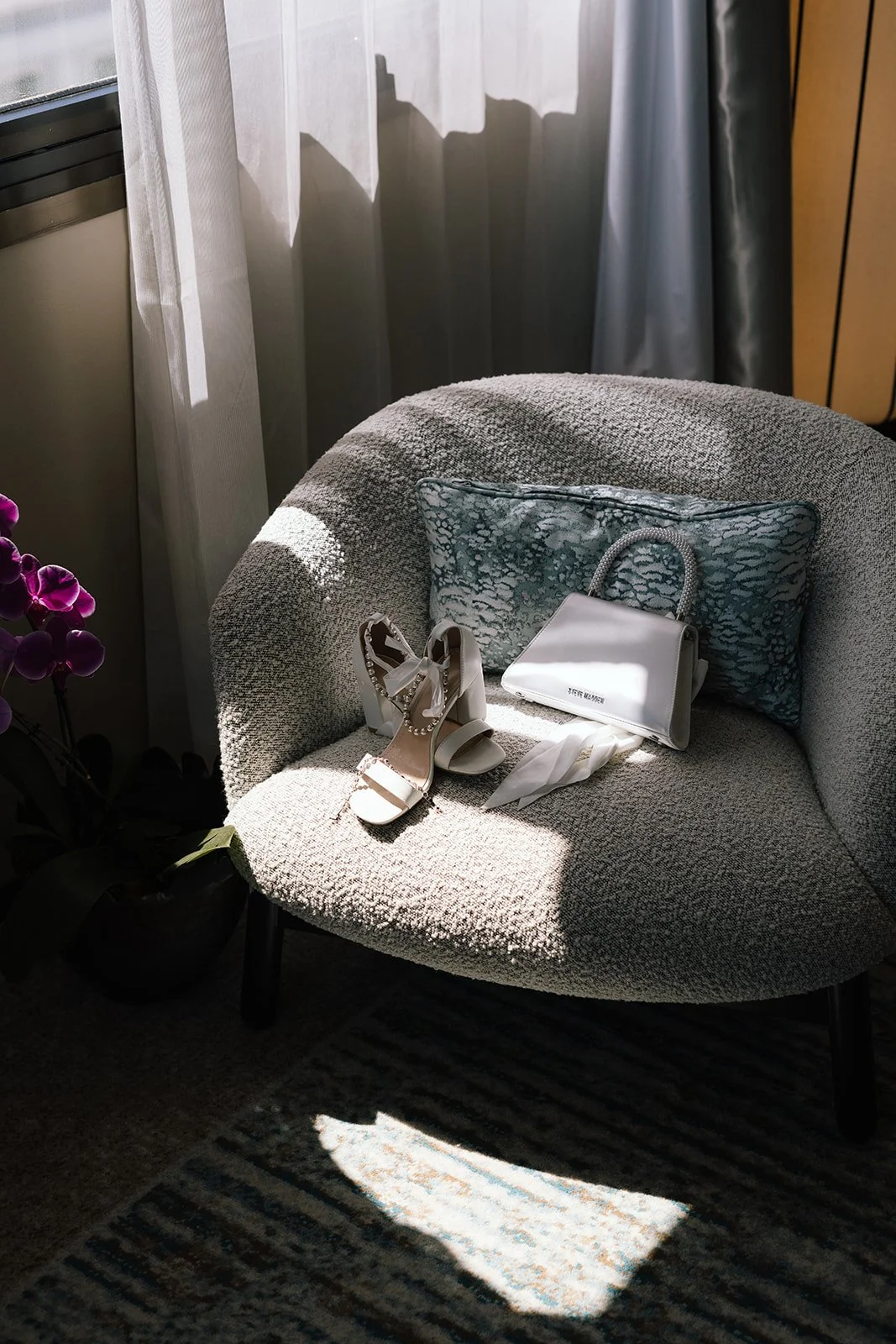 Close-up detail shot of white block heel bridal shoes with pearl ankle straps and a white Steve Madden handbag resting in dramatic sunlight on a textured bouclé armchair at Pan Pacific Perth, photographed by James Di Stefano Photo and Video.
