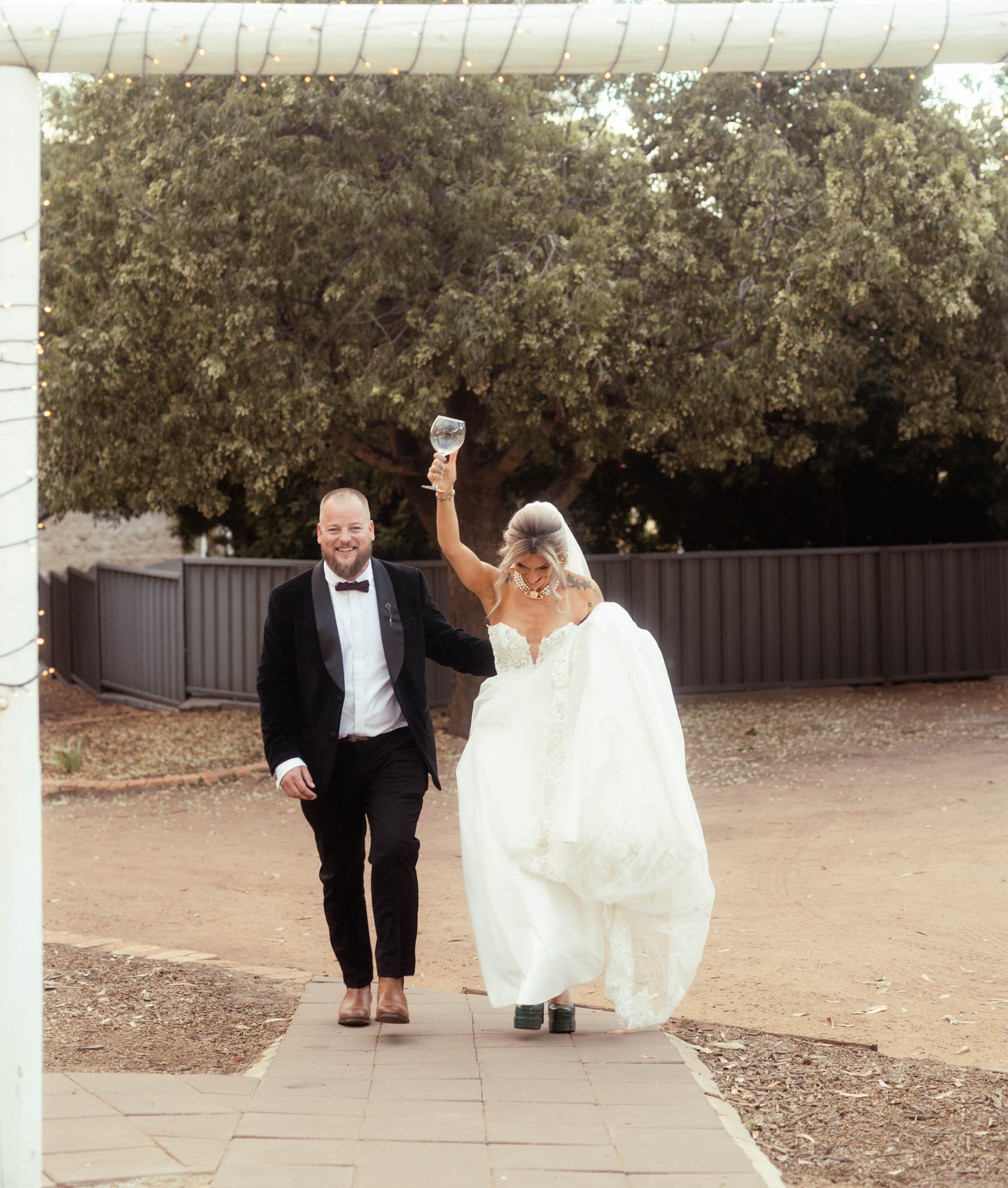 A joyful candid photograph showing the smiling bride and groom walking together along a pathway at Buckland Estate, a hidden outback wedding venue near Perth. The bride holds a celebratory drink while both relax after the ceremony