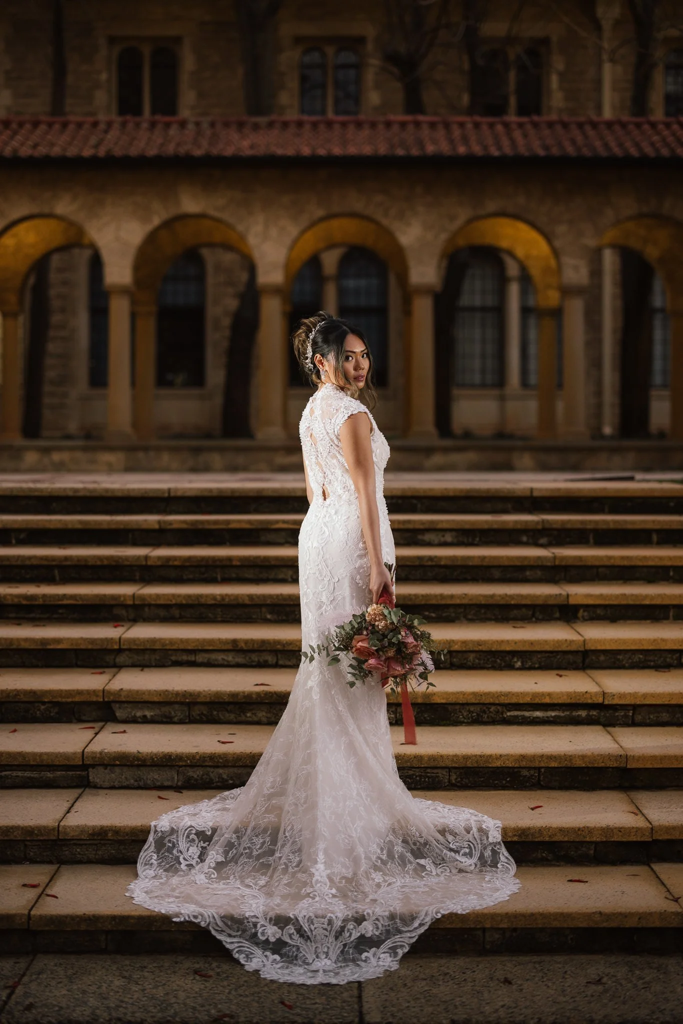 Bride in an intricate lace wedding gown with a sweeping train glances back on sandstone steps before a grand arched colonnade