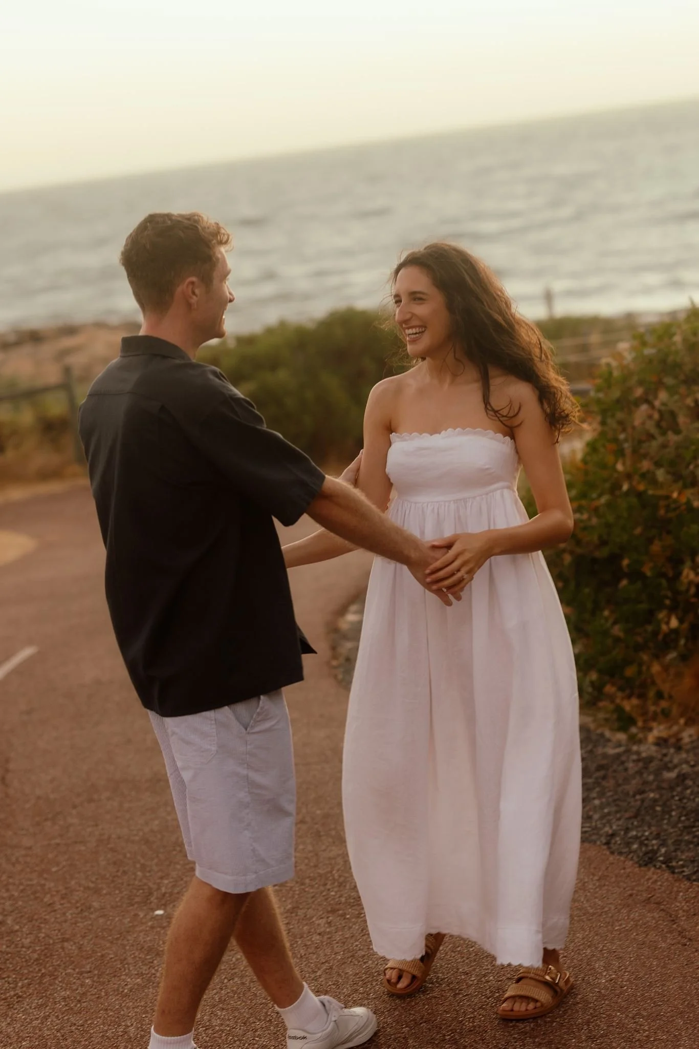 Laughing couple hold hands on a sun-warmed coastal path at Perry's Paddock during a romantic pre-wedding shoot at golden hour