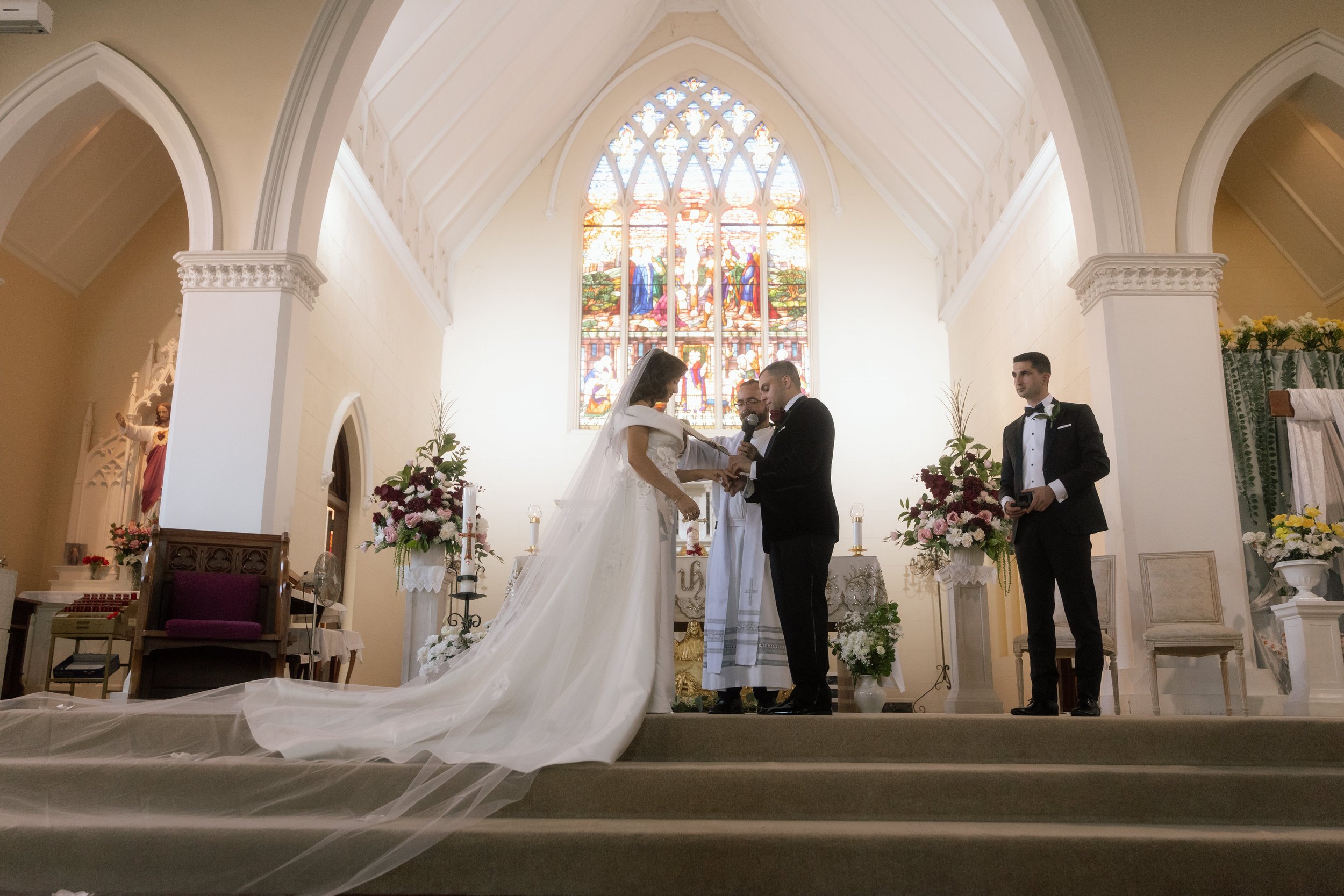 Couple exchanging rings before a priest beneath a soaring Gothic stained-glass window during a cinematic church wedding ceremony